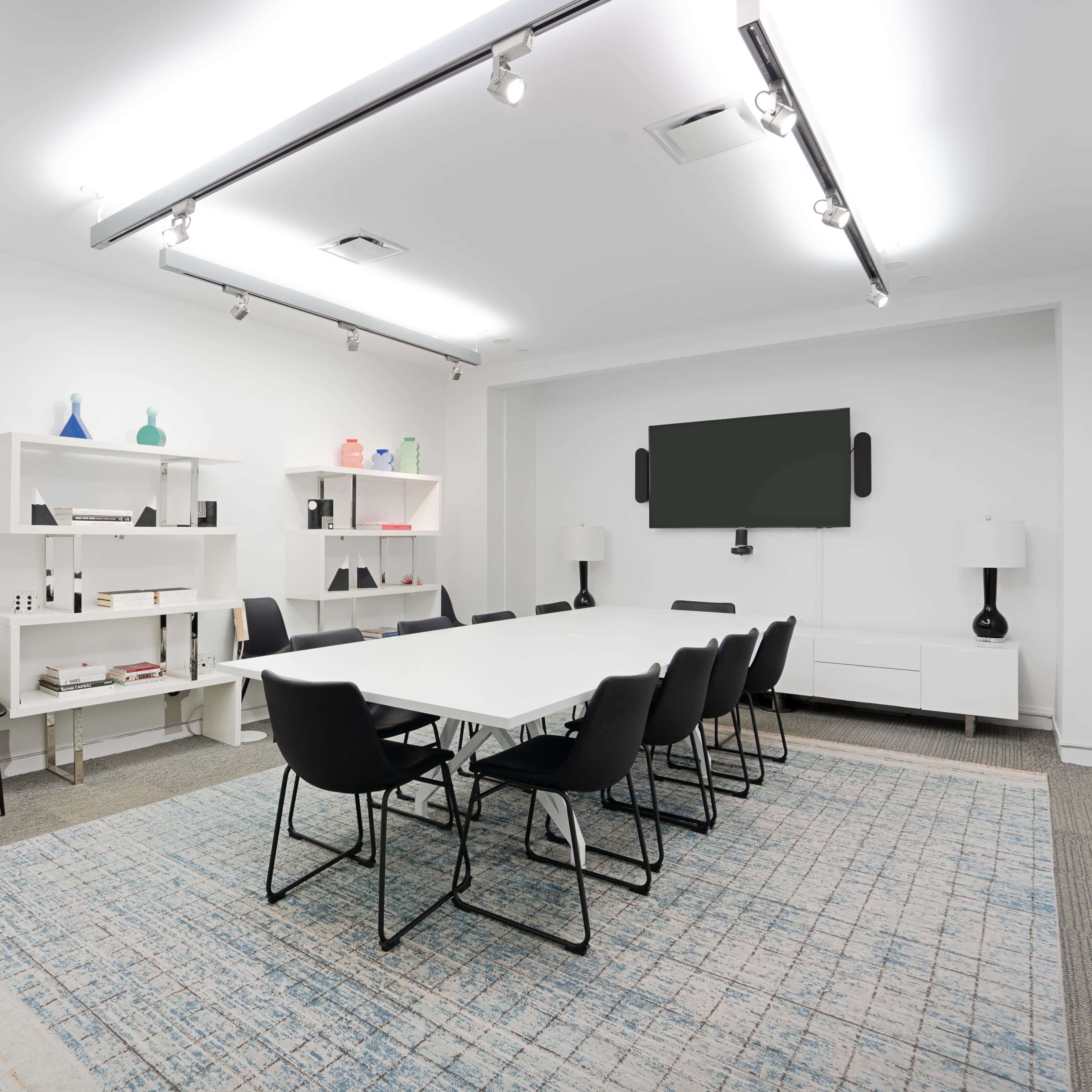 A modern meeting room features a long white table surrounded by black chairs, with shelving and a television on the walls.