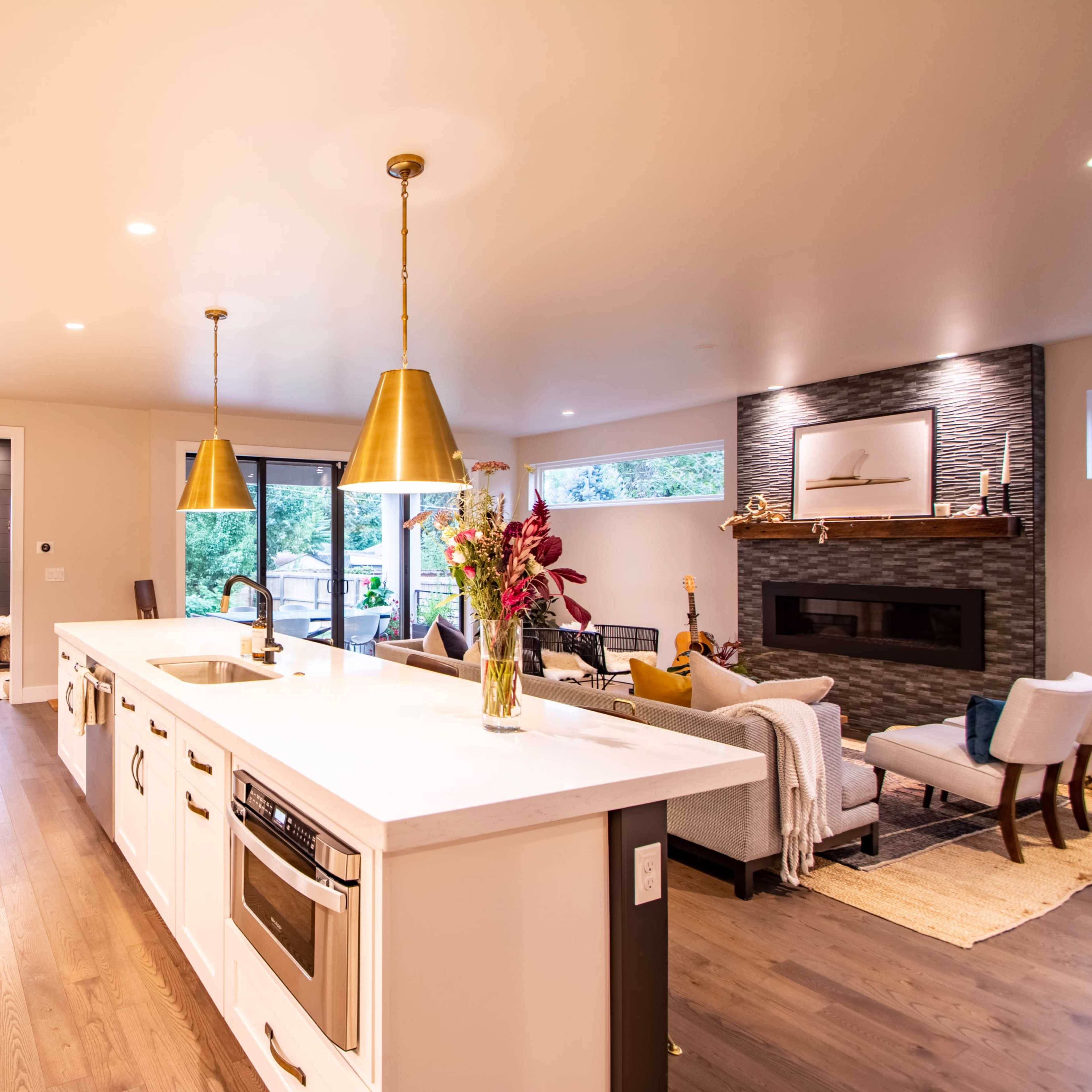 A modern kitchen features white cabinetry, a large island with bar seating, and gold pendant lights, adjoining a living area with a fireplace and large windows.