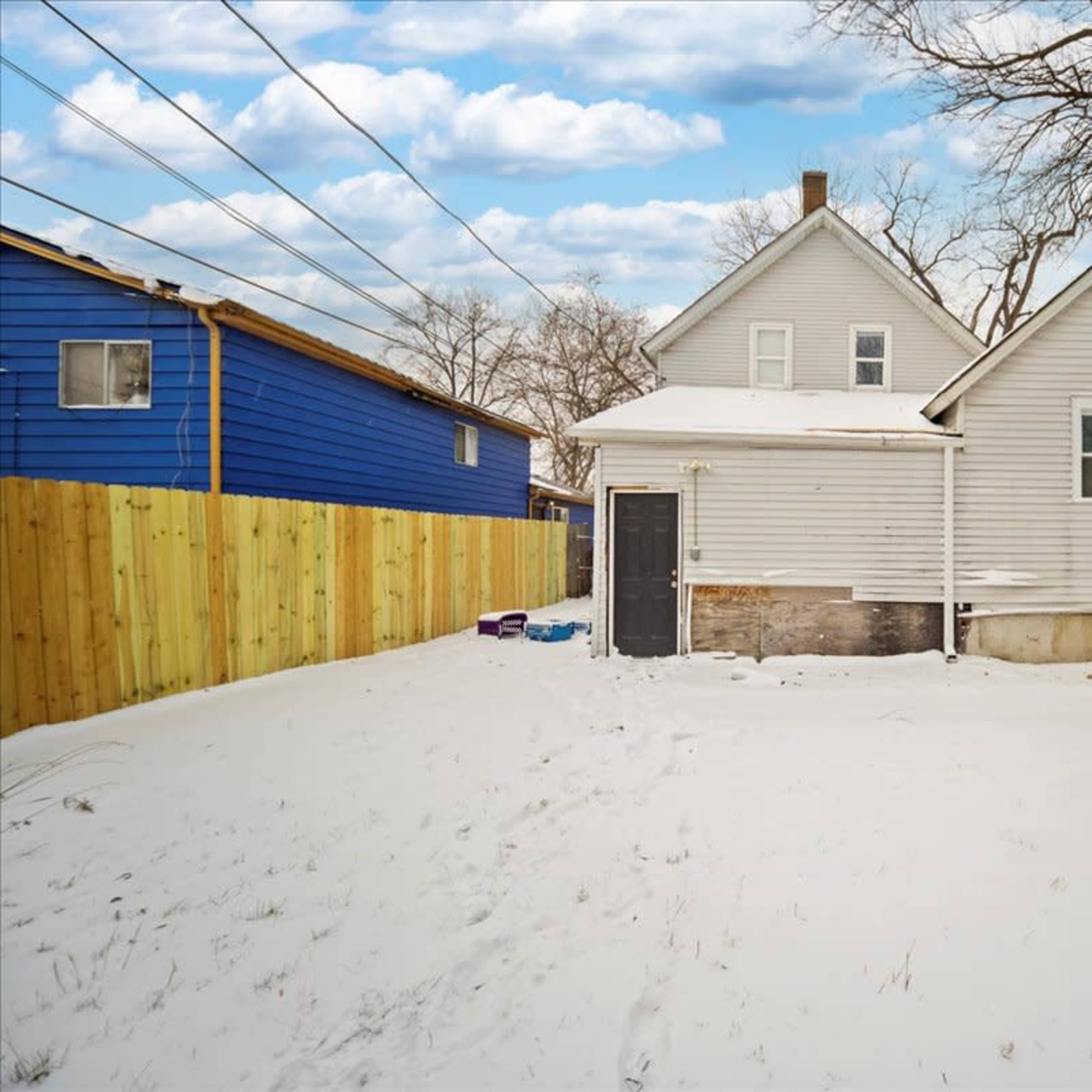 A two-story house with a gray exterior is surrounded by snow and a wooden fence.
