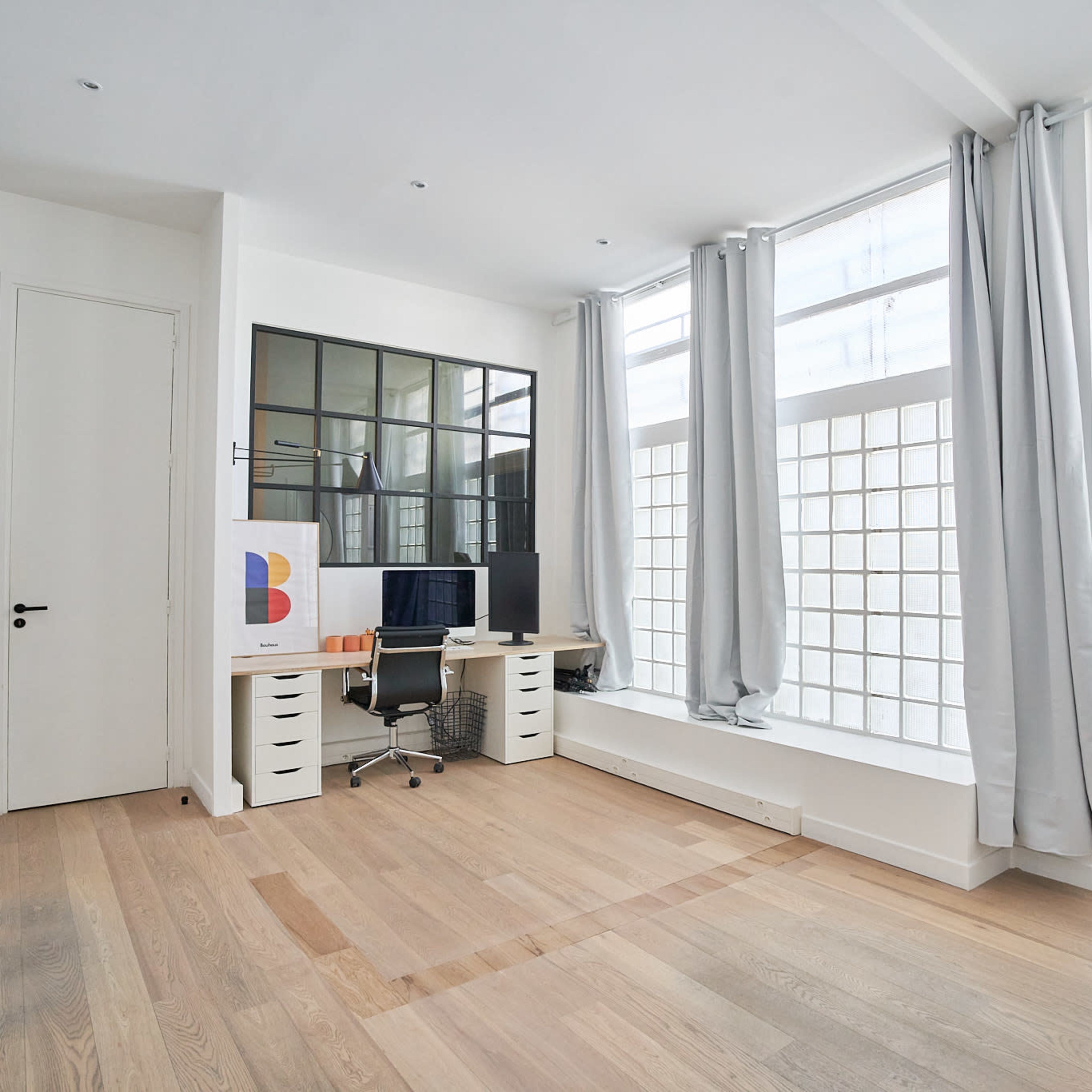 A modern office space with a desk and computer, large glass block windows, and light-colored wooden flooring.