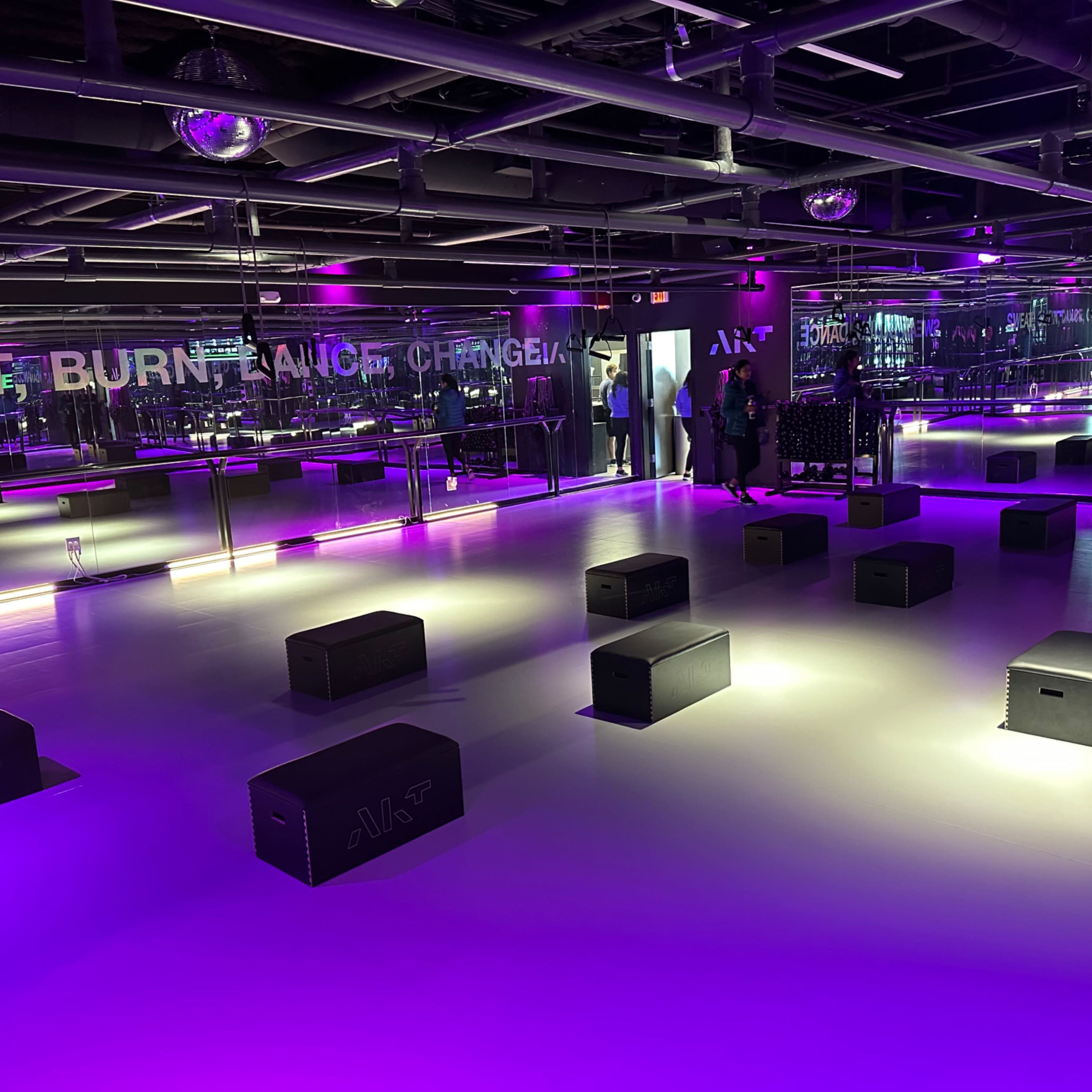 A modern fitness studio with a mirrored wall, purple lighting, and black exercise boxes arranged on the floor.