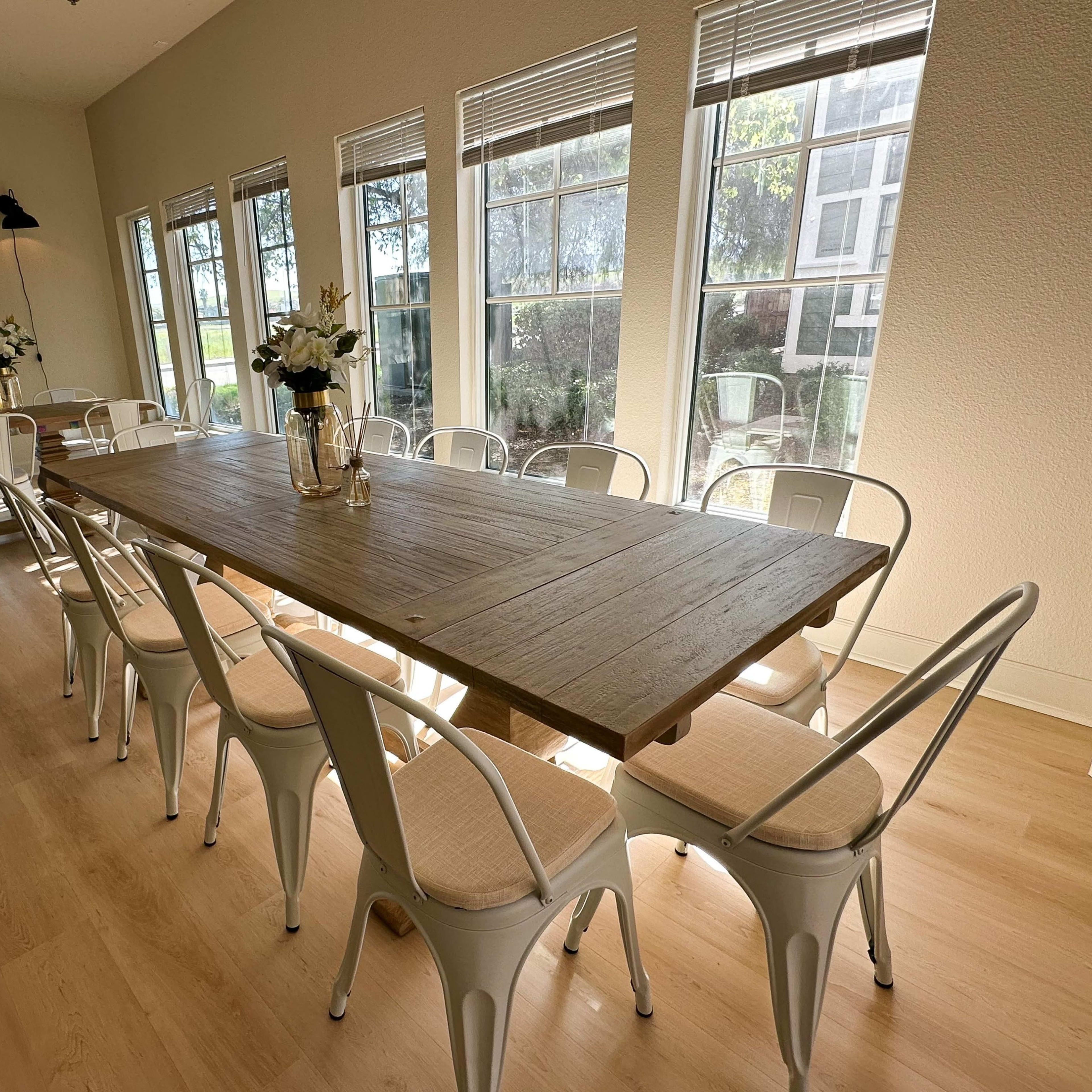 A long wooden table surrounded by white chairs is set in a brightly lit room with large windows.