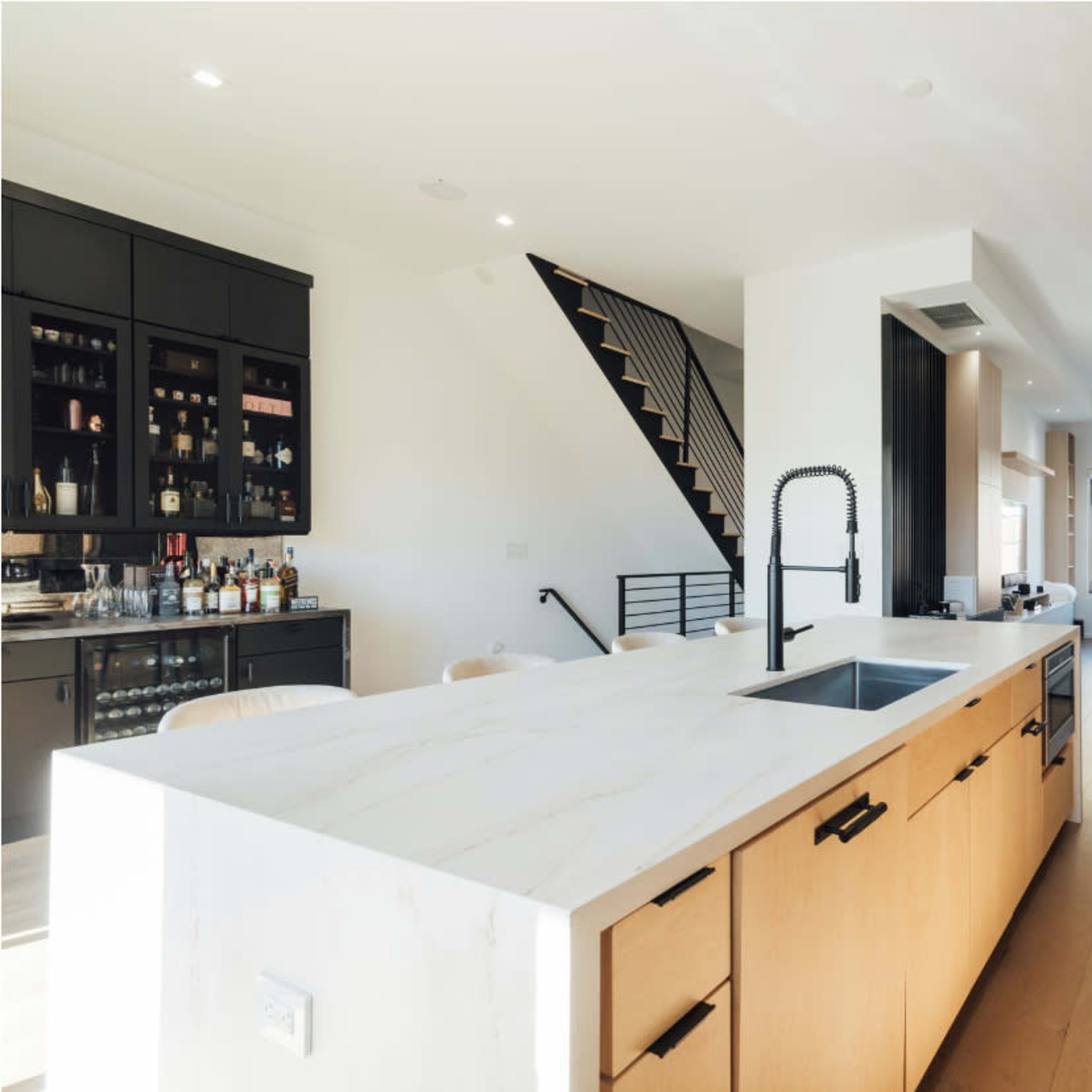 A modern kitchen with a central island, wooden cabinets, and a staircase leading to an upper level, while a living area is visible in the background.