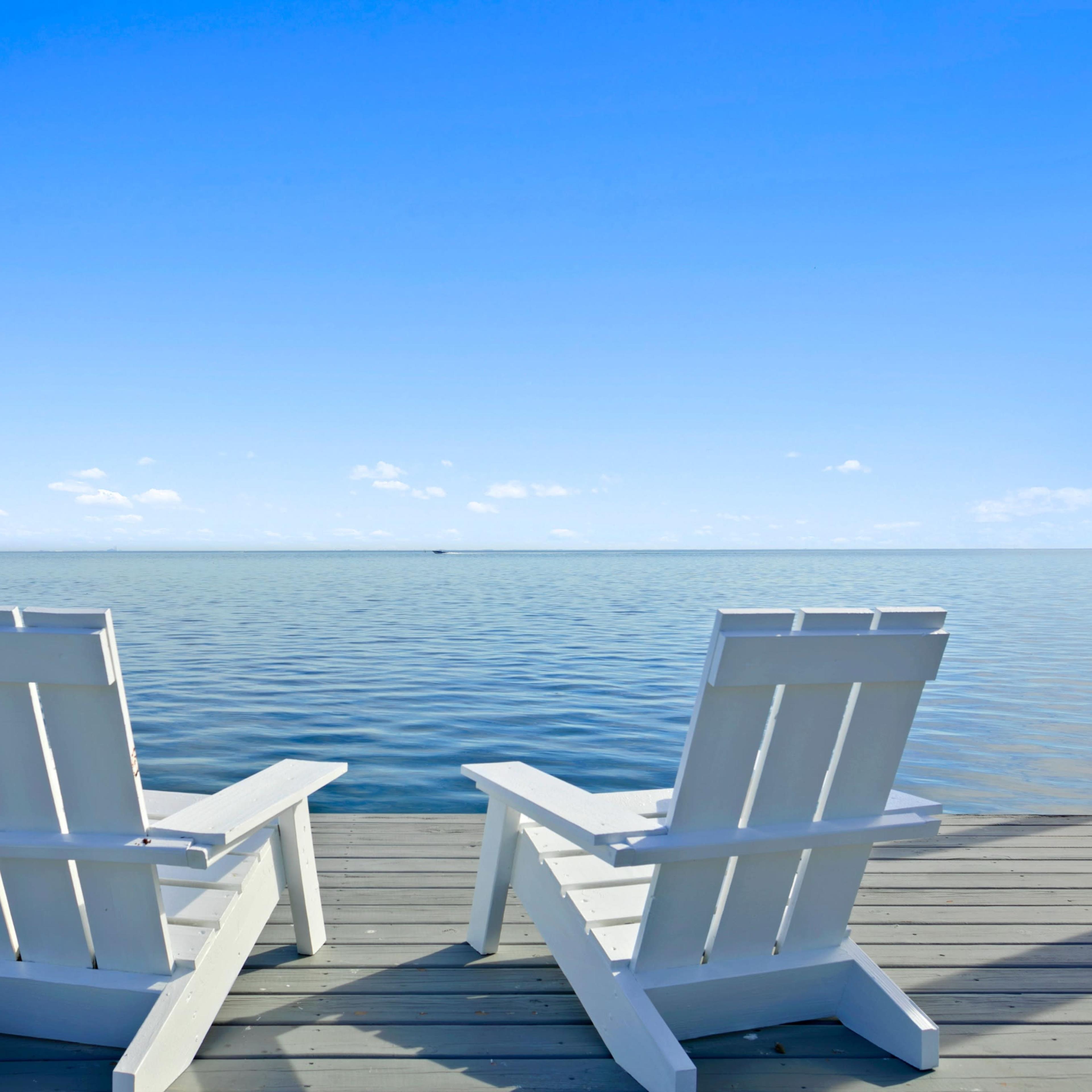Two white Adirondack chairs are positioned on a wooden dock overlooking a calm body of water under a clear blue sky.