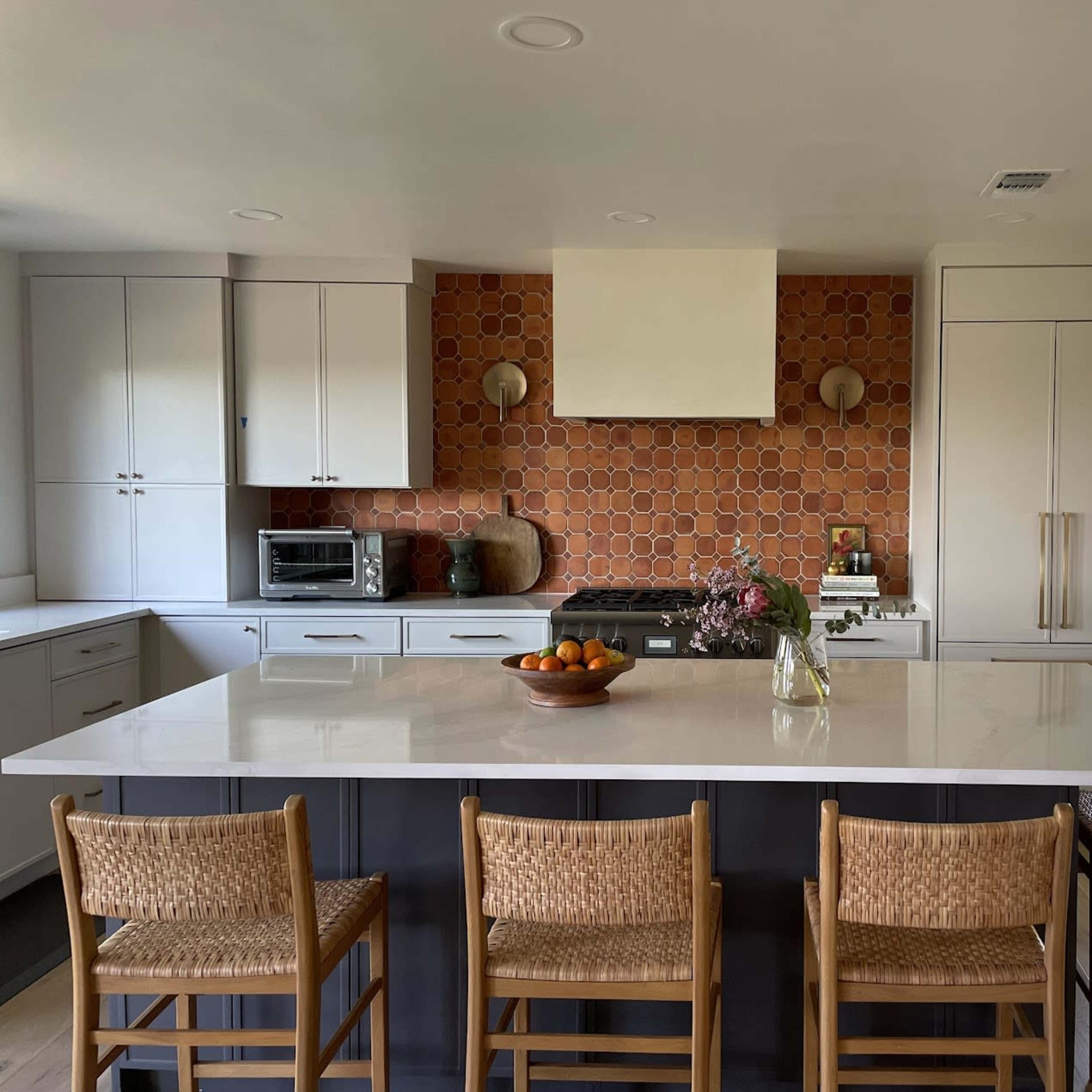 A modern kitchen features a large island with wooden stools, a hexagonal tile backsplash, and a view of an outdoor space through a window.