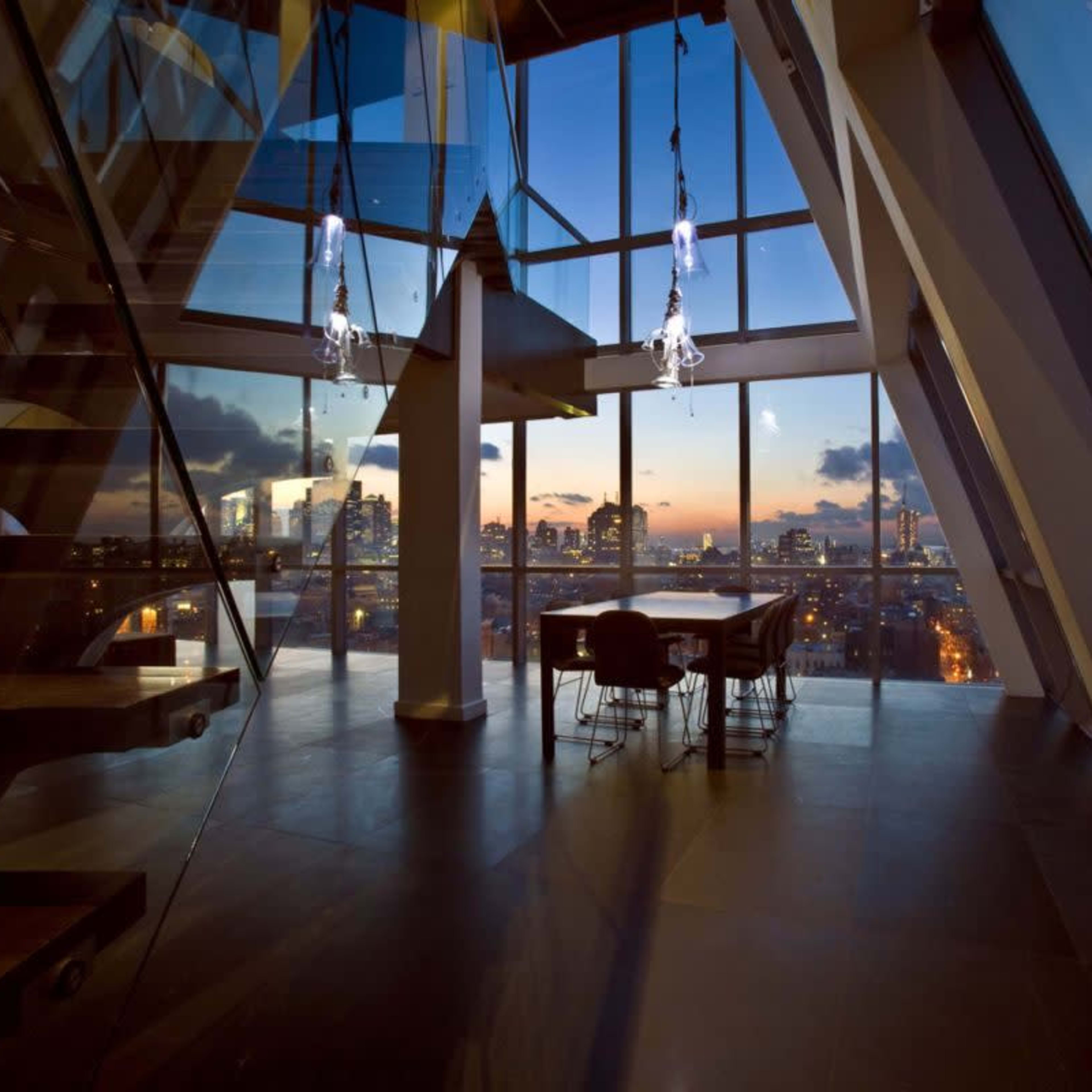 A modern interior features a staircase leading to a dining area with large windows showcasing a city skyline at dusk.