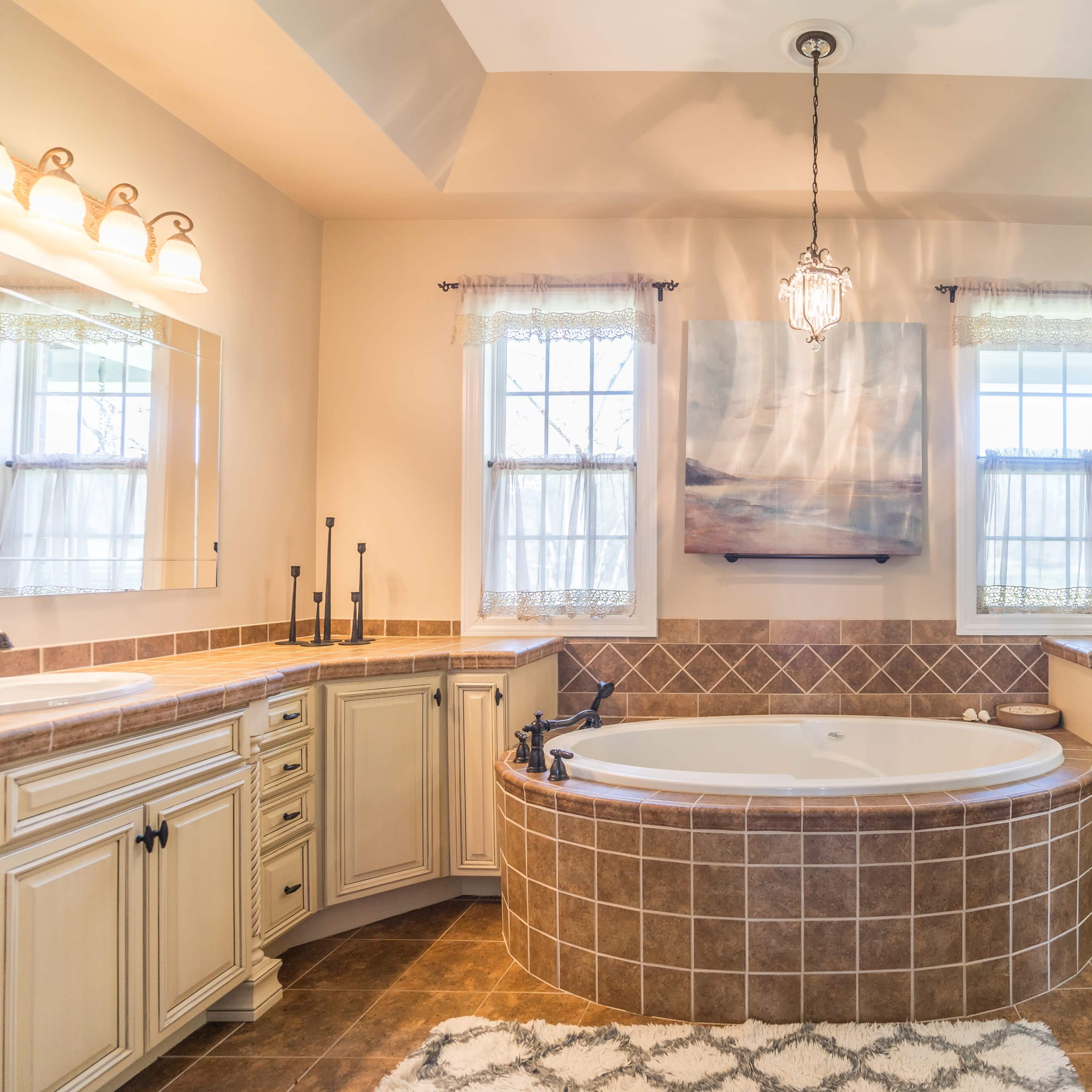 A spacious bathroom featuring a large oval bathtub surrounded by beige cabinetry and two windows with sheer curtains.