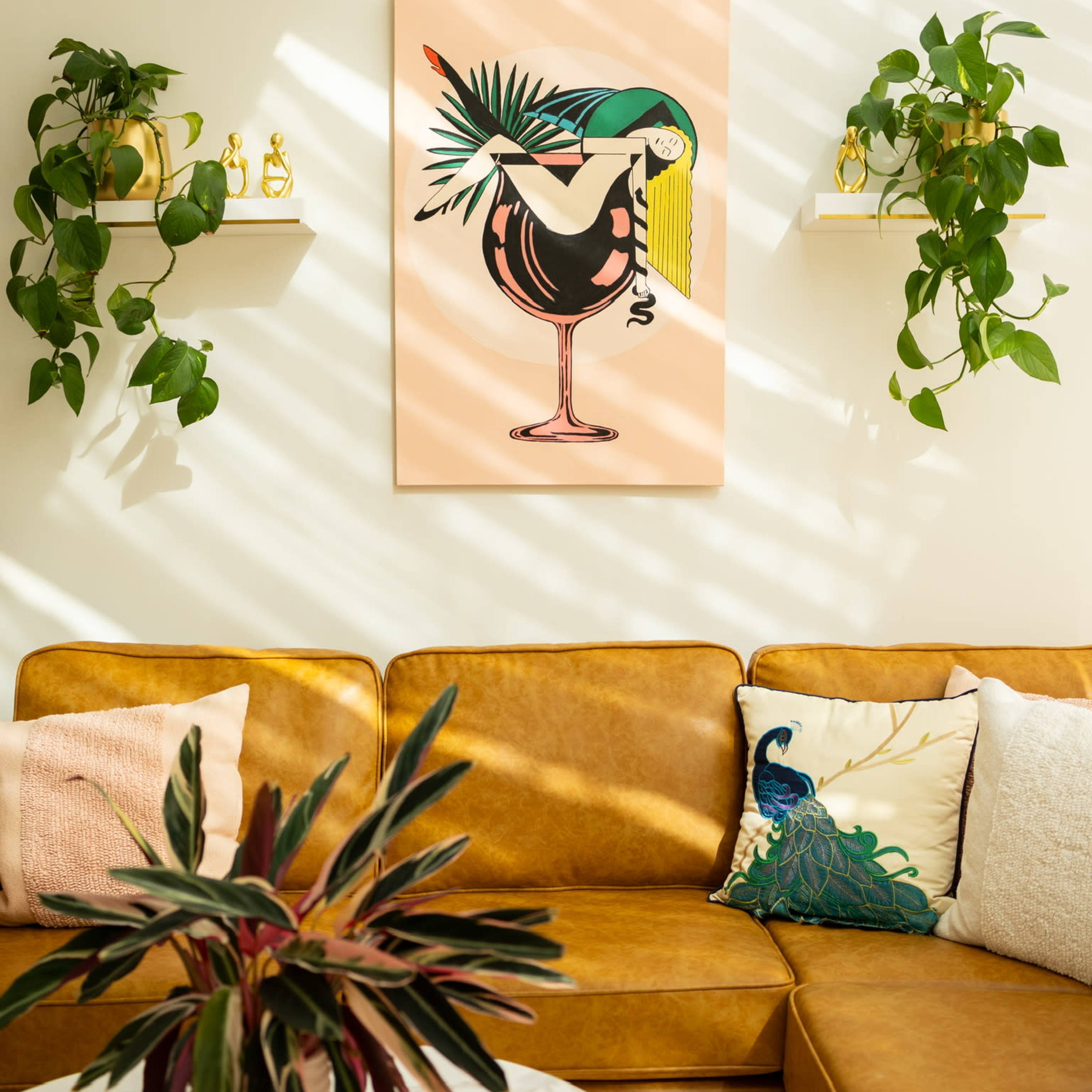 A living room with a brown leather couch, a vibrant artwork featuring a bird and cocktail, and green plants on shelves.