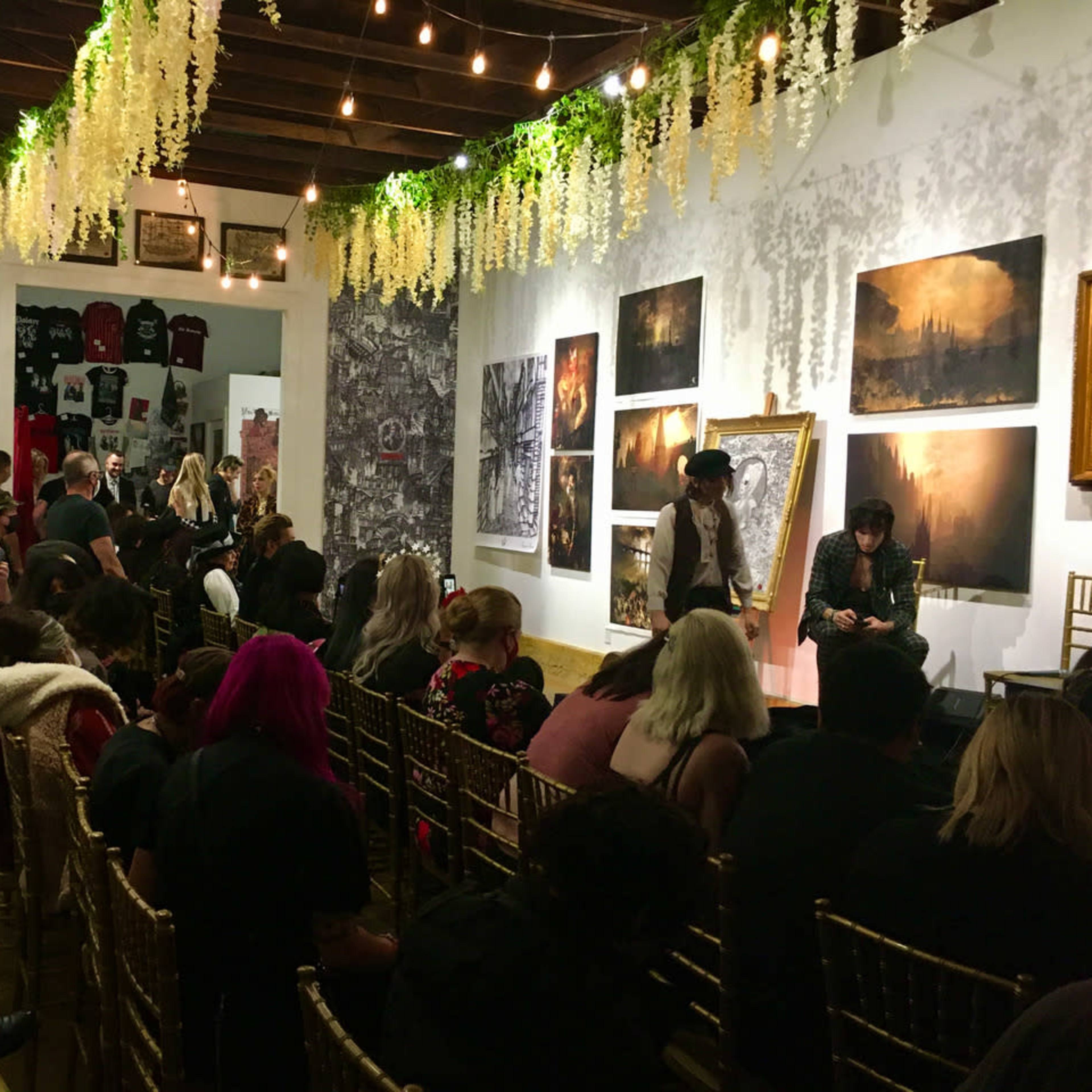 A crowd of people seated in a gallery space adorned with artwork on the walls and decorative hanging plants overhead.