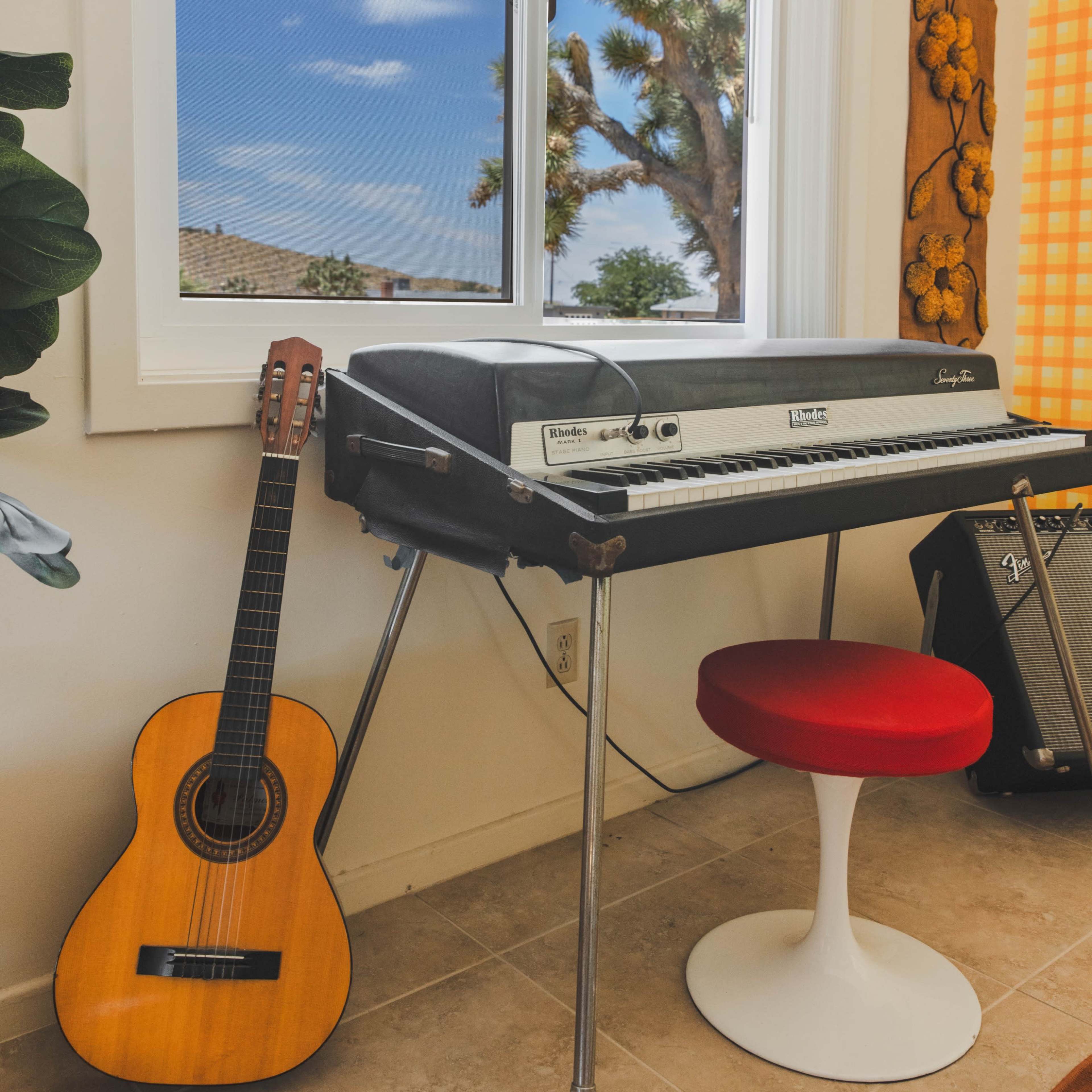 A guitar leans against a keyboard setup near a window overlooking a desert landscape.