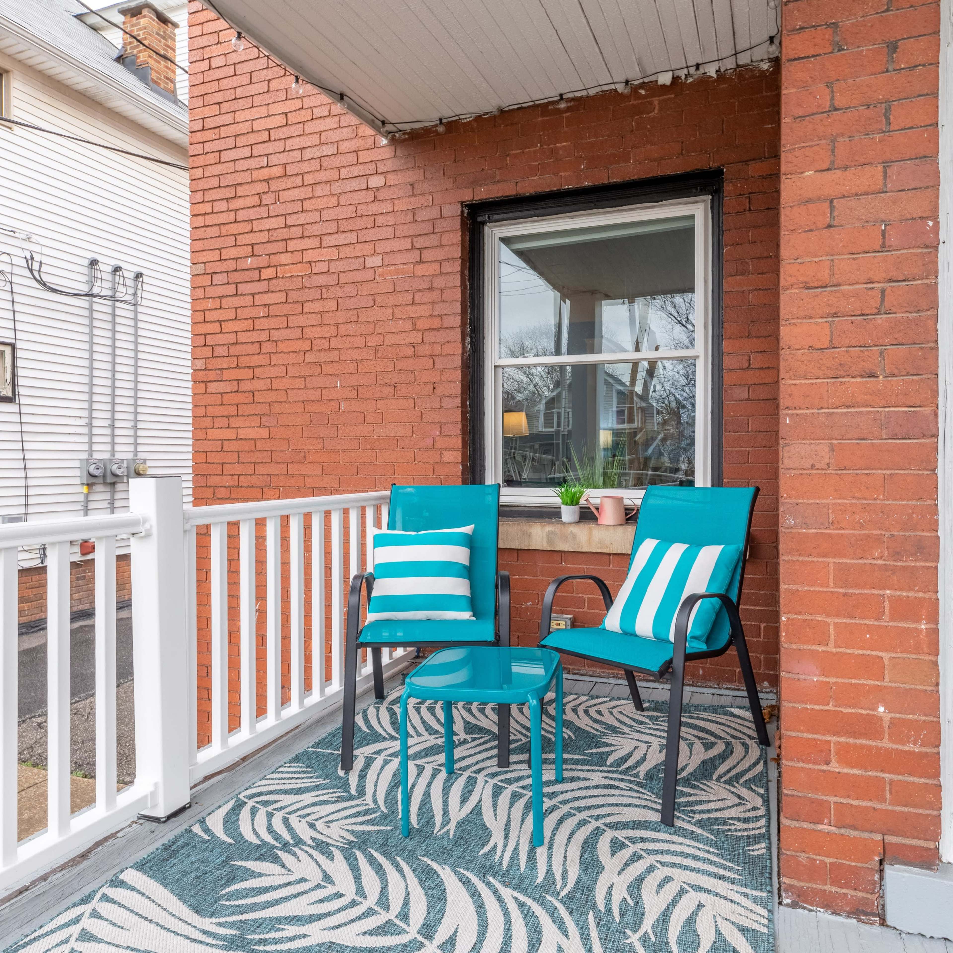 A small porch features two teal chairs and a matching table on a patterned rug, with a window adjacent to a front door.