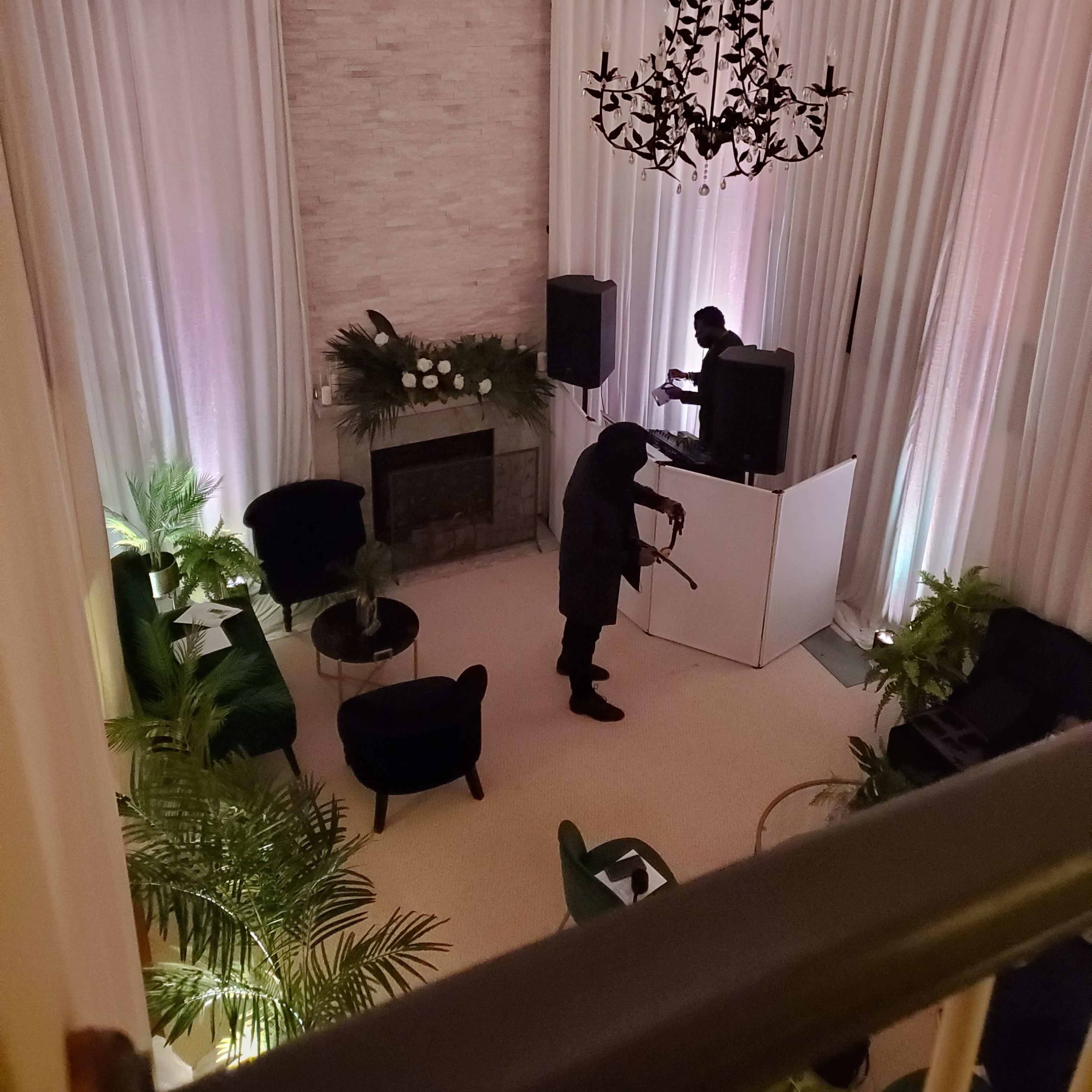 A person plays a violin in a spacious room with a fireplace, large curtains, and green plants, while another individual operates equipment at a DJ booth.