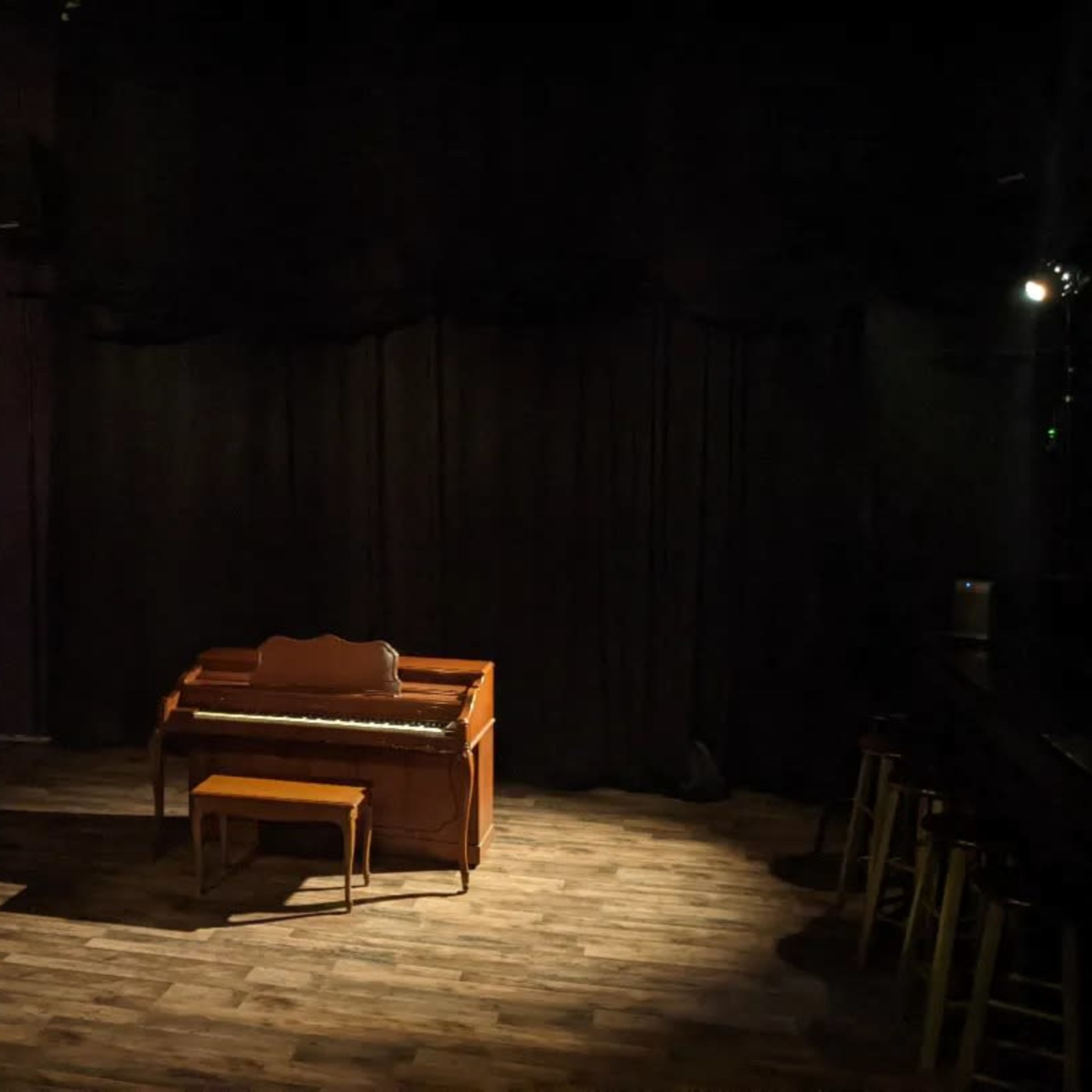 A dimly lit stage with a wooden piano and a small chair set against a backdrop of black curtains.