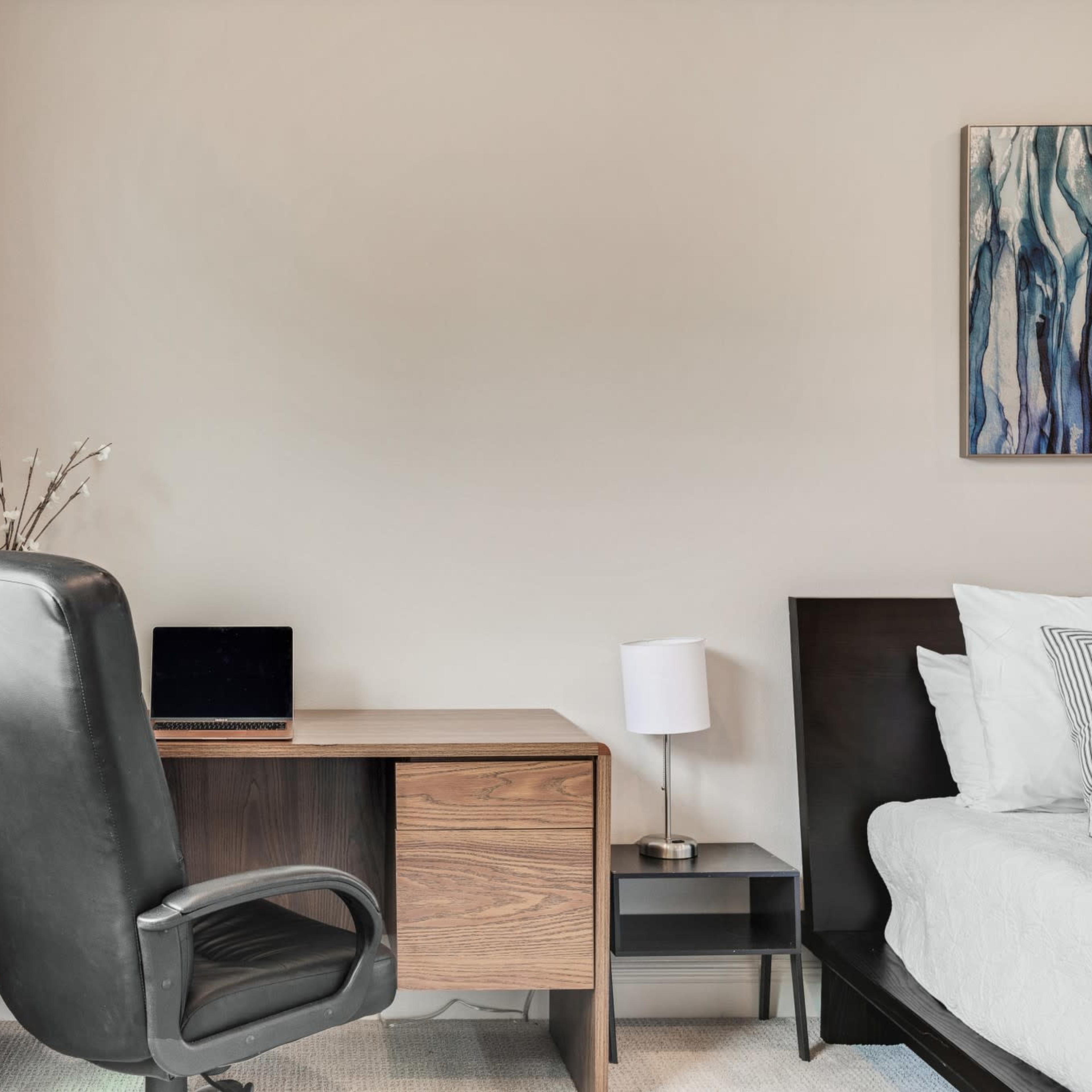 A minimalistic room featuring a wooden desk with a laptop, a black swivel chair, and a bed, along with a bedside lamp and a window with sheer curtains.