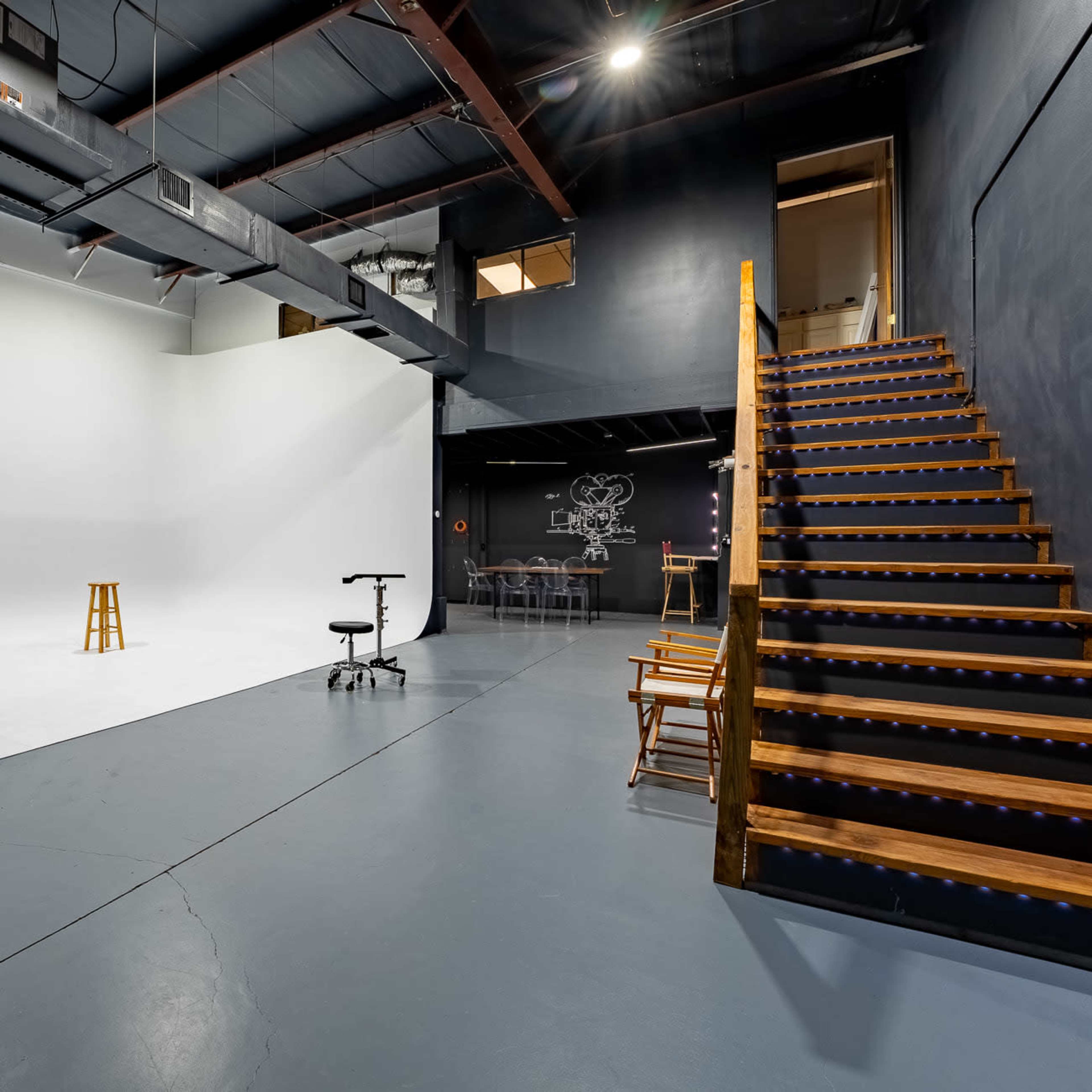 A spacious studio with a white backdrop, wooden stairs leading to an upper level, and various furniture arranged for shooting or meetings.