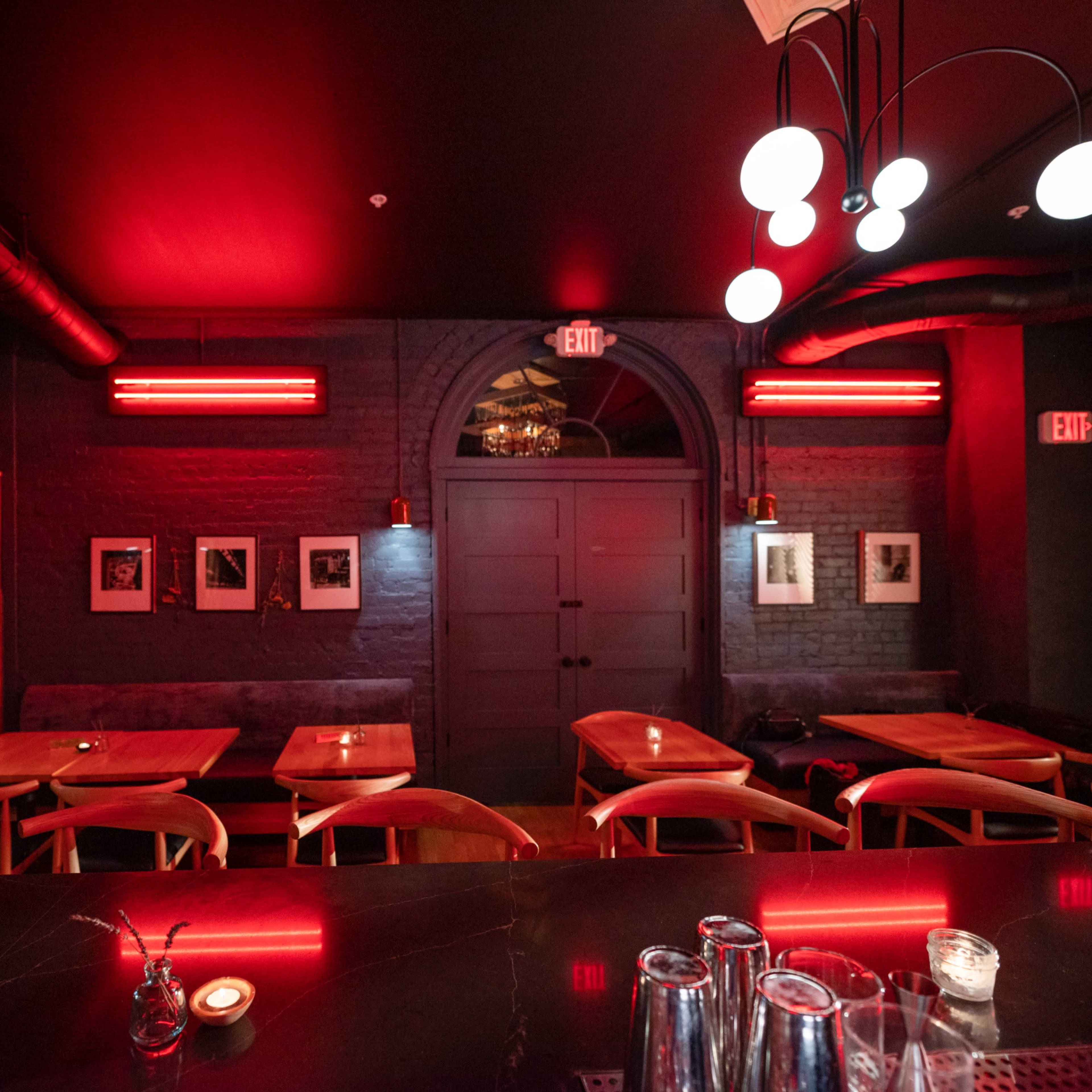 A dimly lit restaurant interior with red lighting, featuring wooden tables, framed photographs on the walls, and a bar area with glasses.
