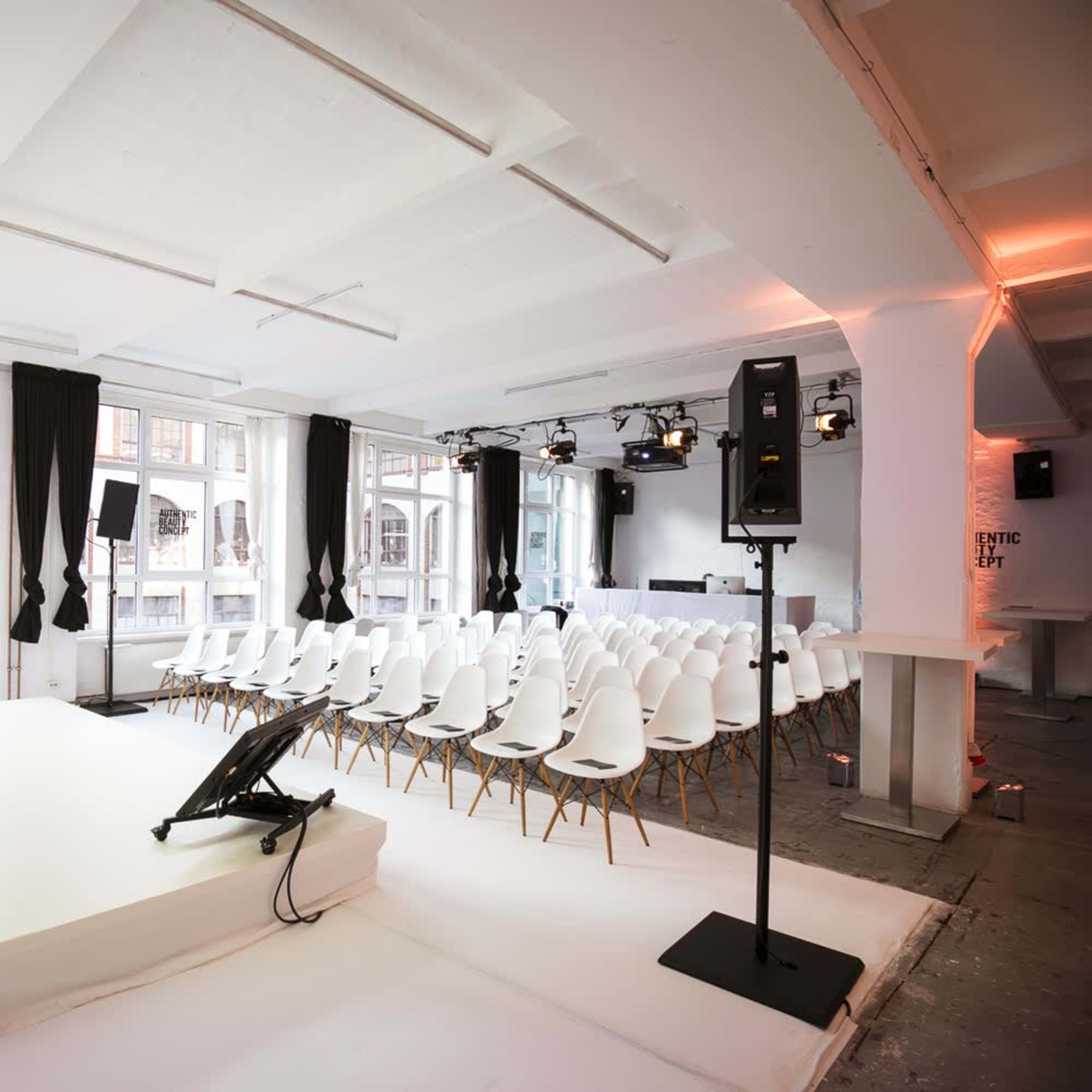 A minimalist event space set up with a stage, a row of white chairs, and large windows letting in natural light.