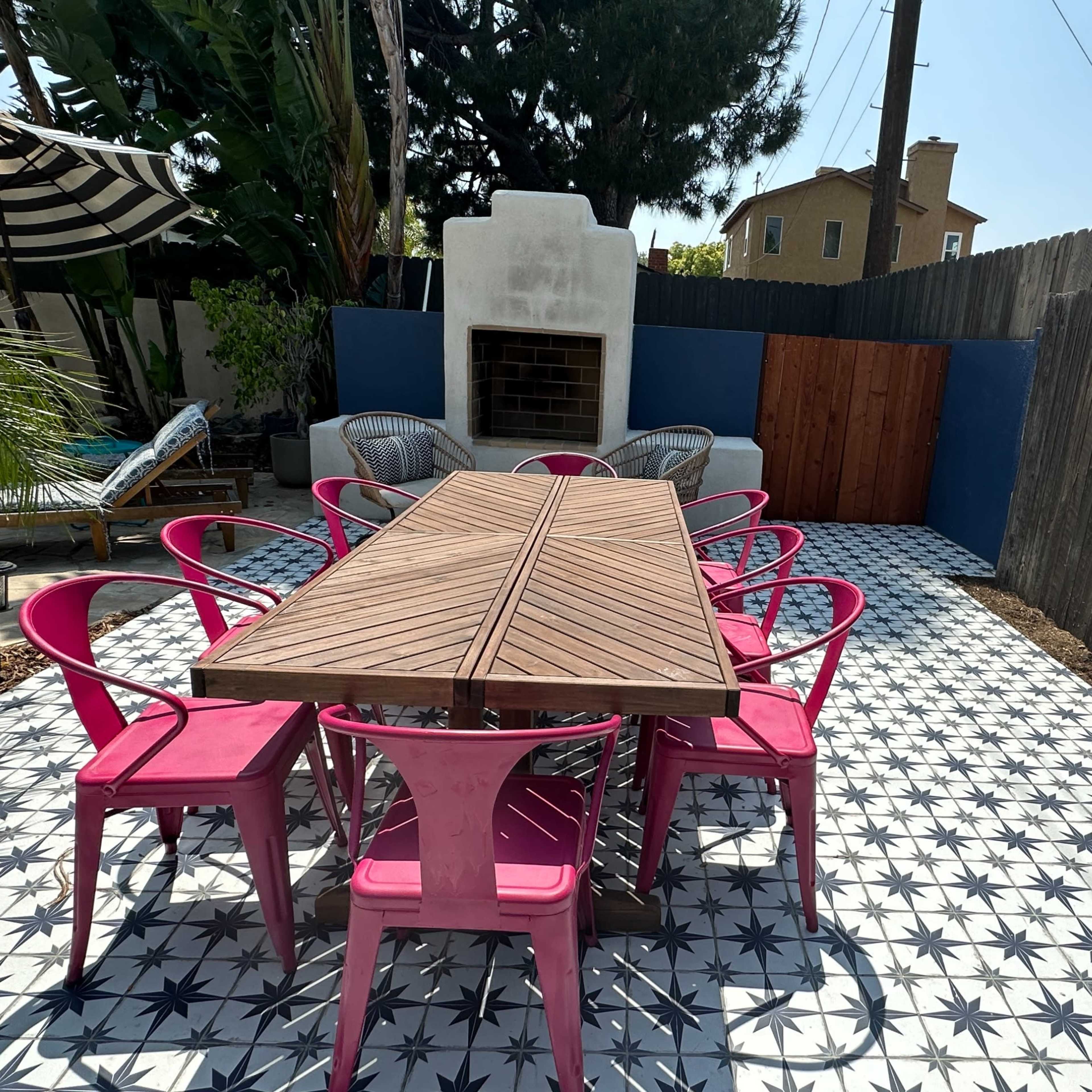 A long wooden table surrounded by pink chairs is set on a patterned tile patio with a fireplace and a wooden fence in the background.