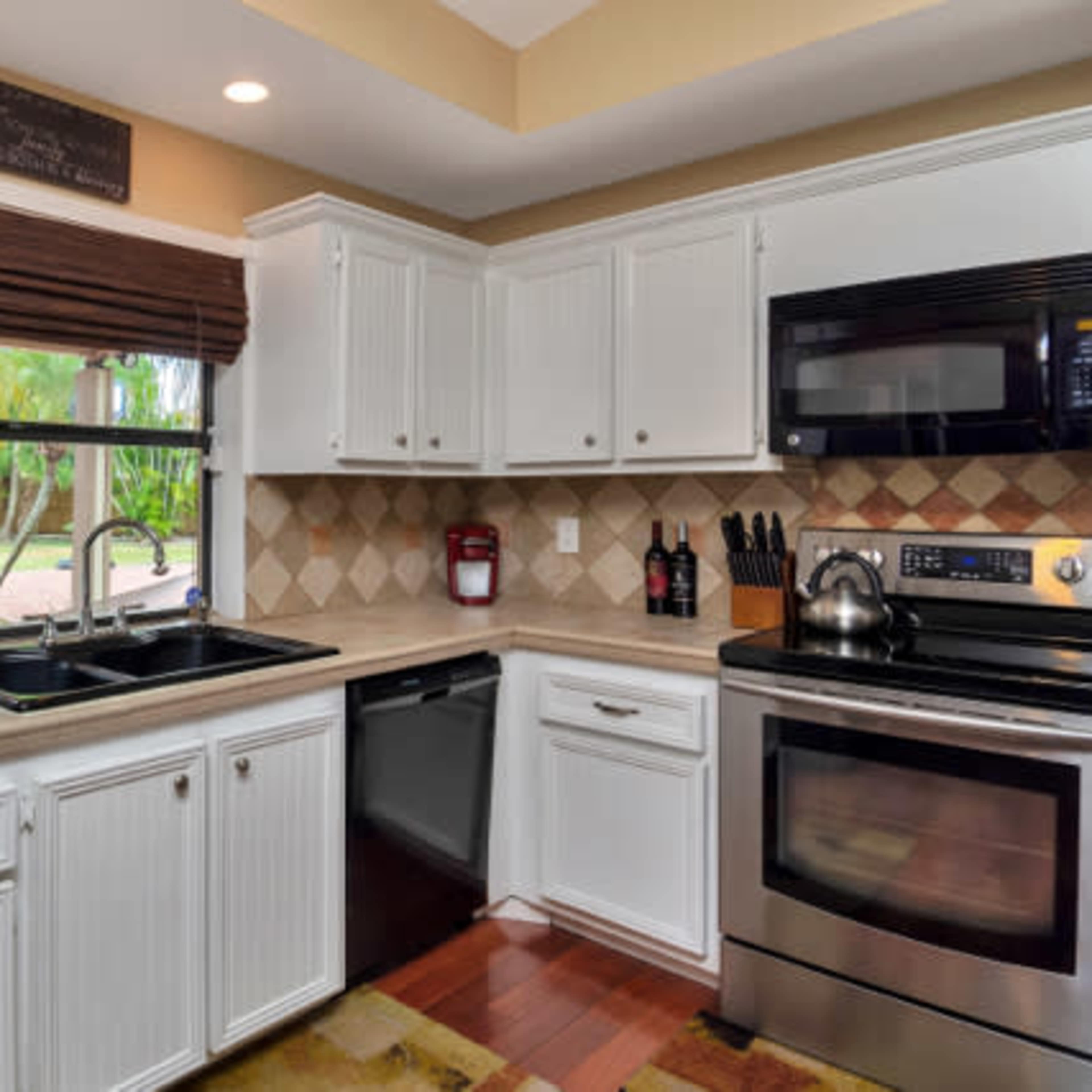 A modern kitchen featuring white cabinetry, a stainless steel refrigerator, a black microwave, and a double sink under a window with a view of greenery outside.