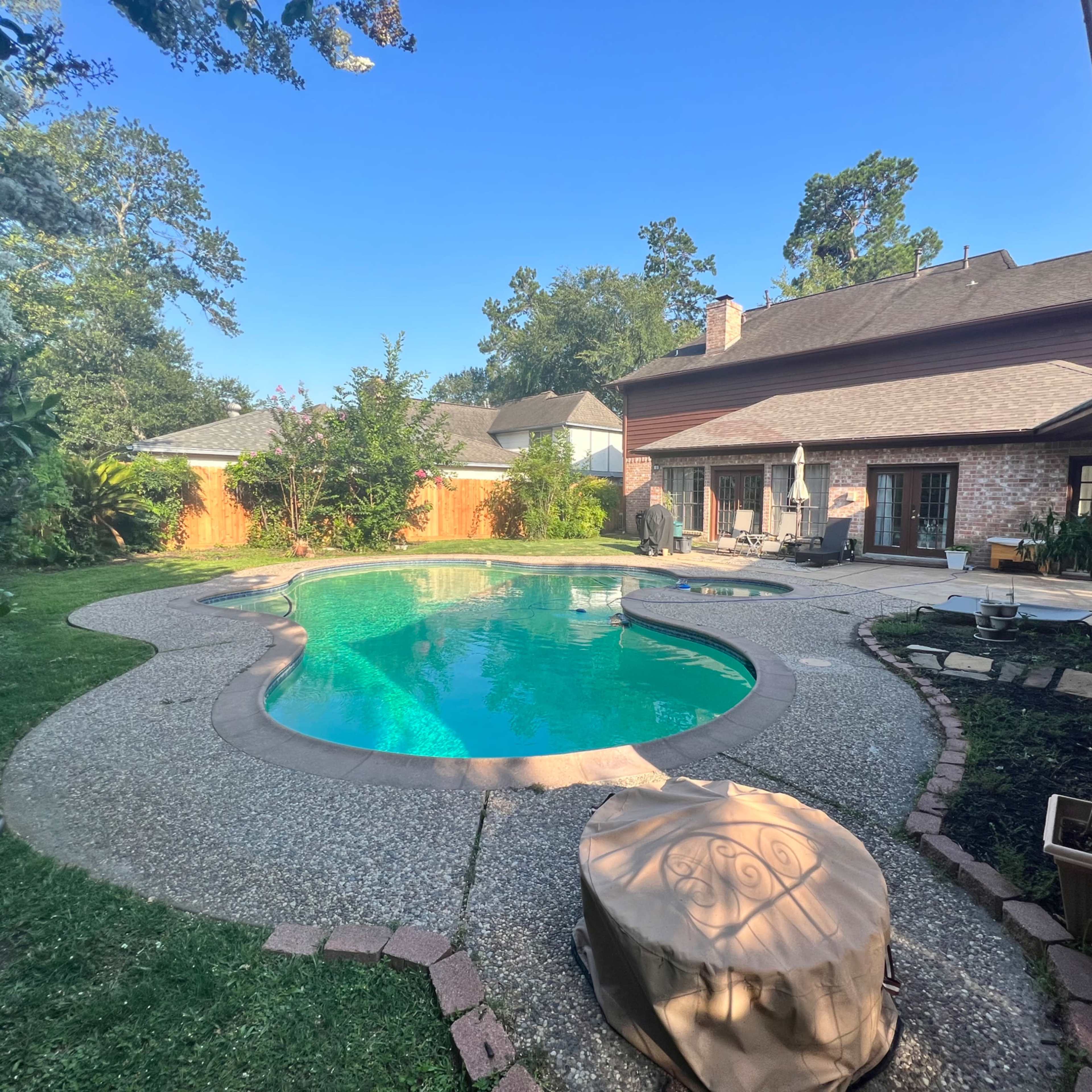 A backyard with a curved swimming pool surrounded by a patio area and trees in the background.