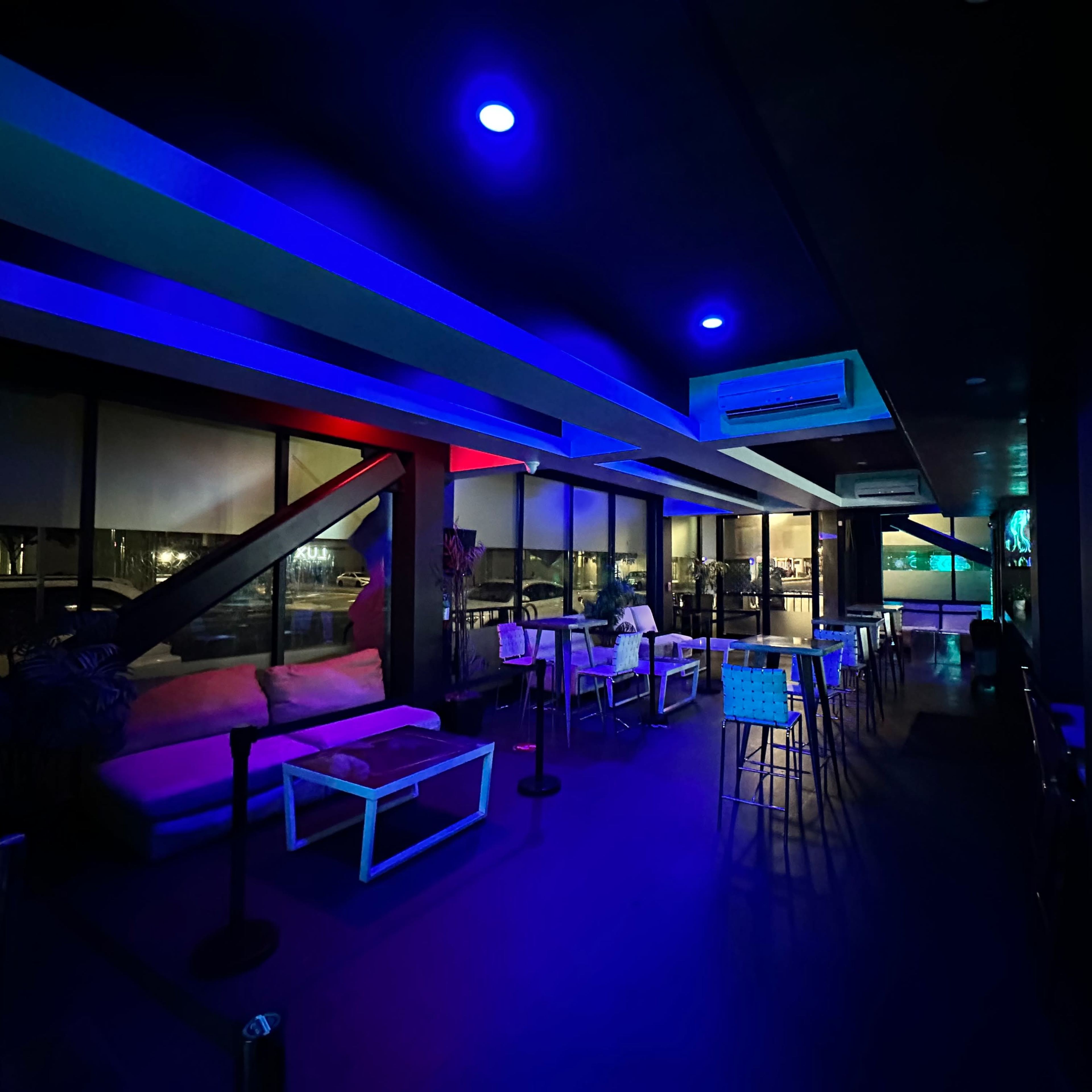 A dimly lit lounge with blue and red lighting, featuring tables, chairs, and large windows overlooking an urban view at night.