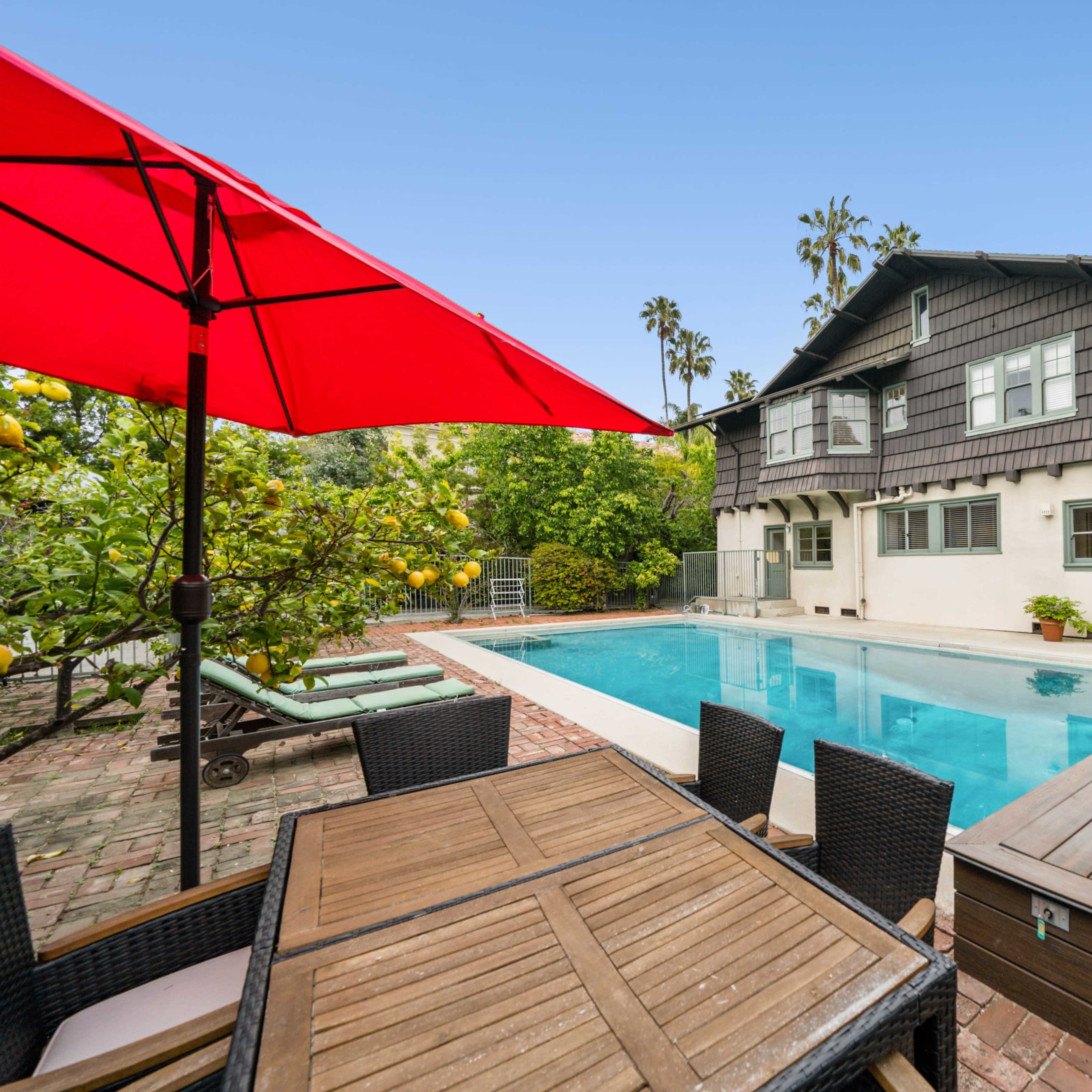 A backyard with a swimming pool, a dining table, and a house surrounded by greenery and palm trees.