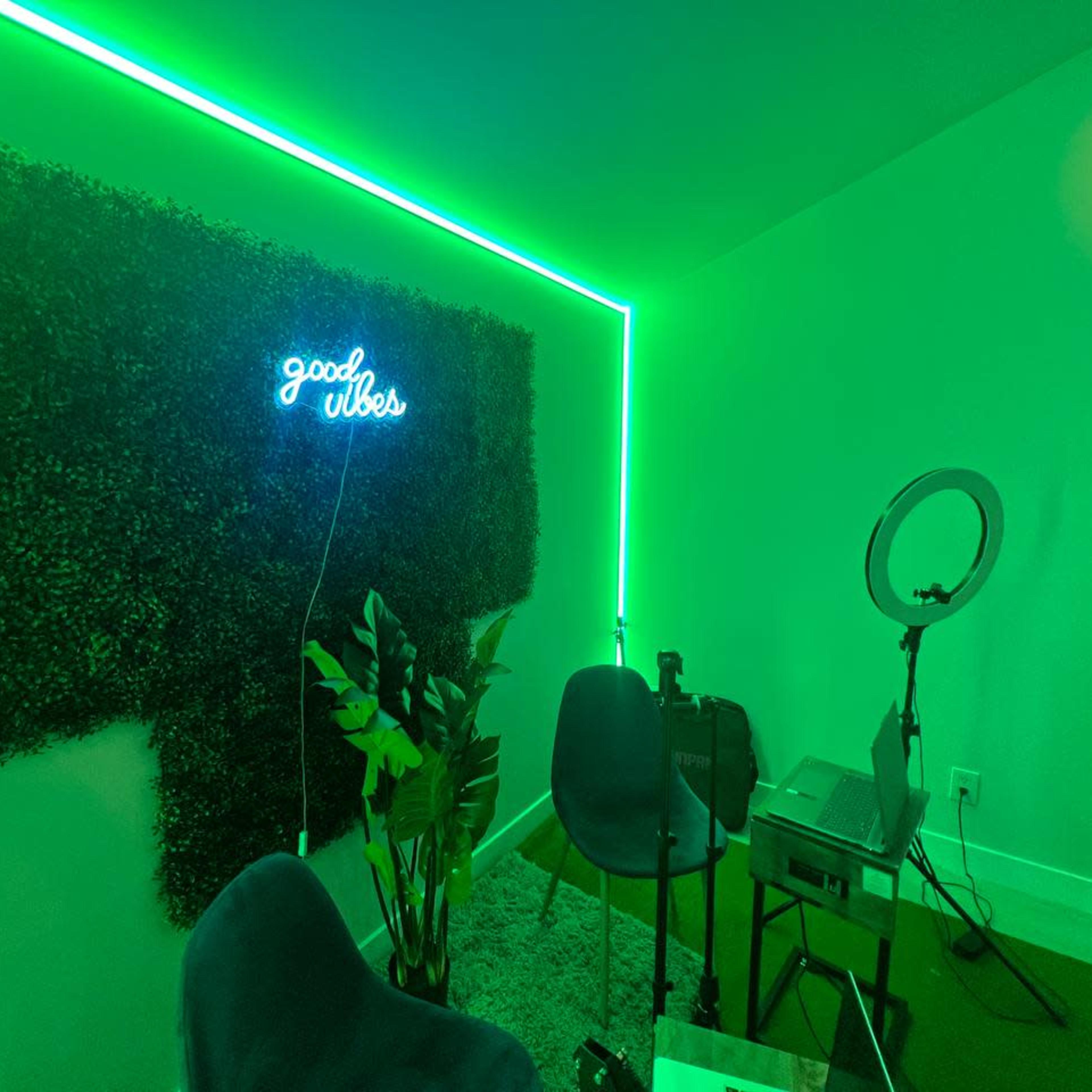 A small room with green neon lighting, featuring a "good vibes" sign, a plant, and a setup for video recording or streaming.