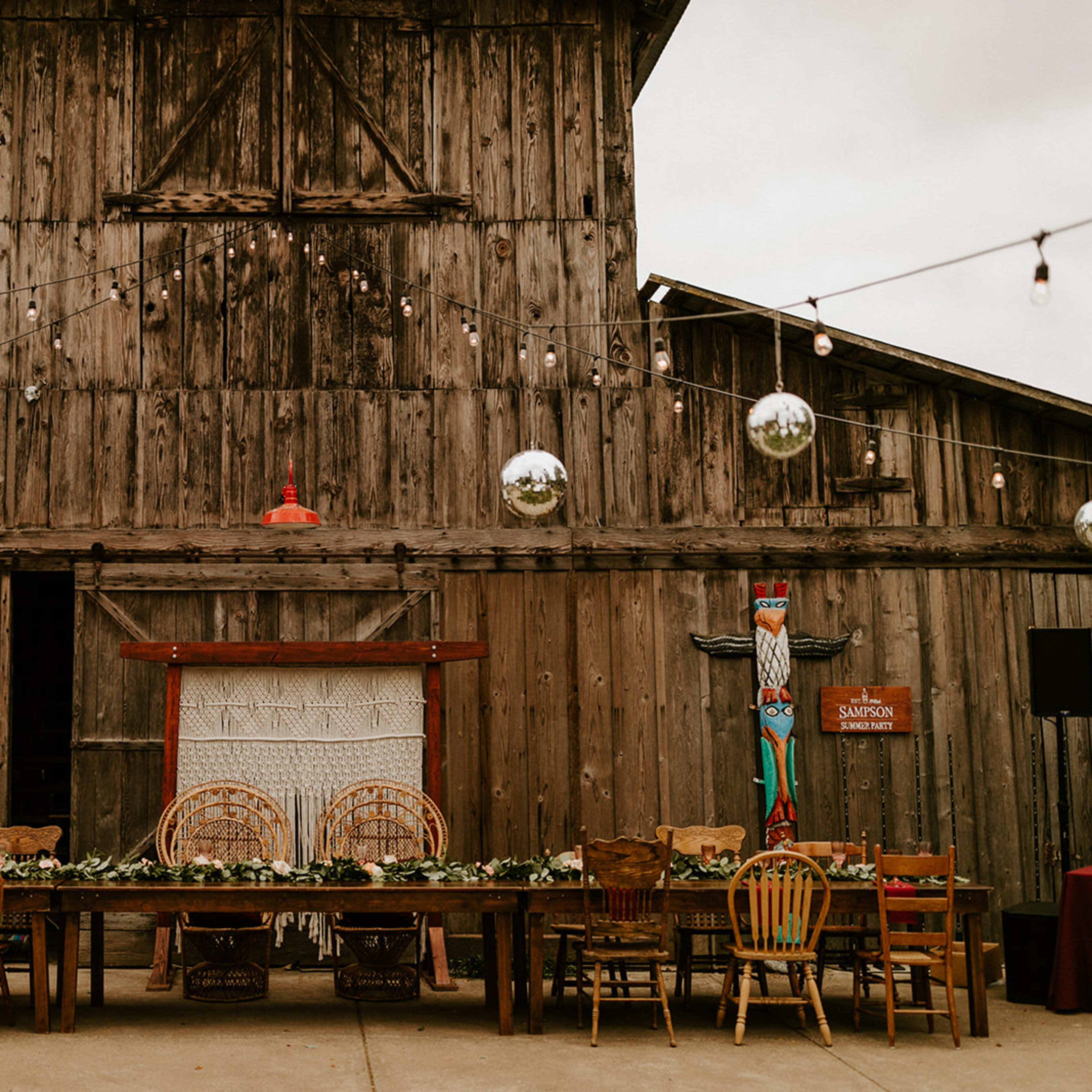 A long dining table is set in front of a rustic wooden barn, adorned with hanging disco balls and string lights.