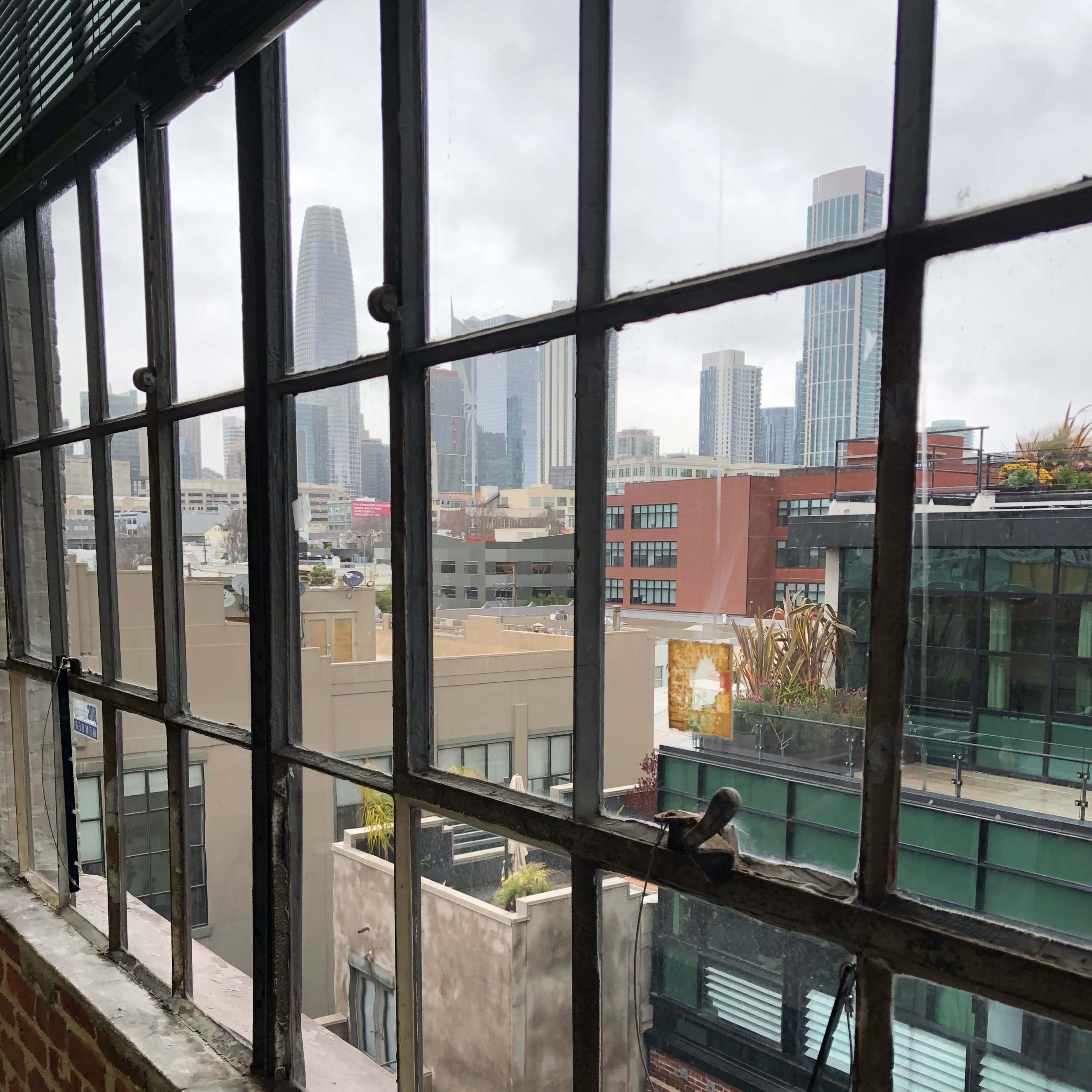 A view from a window with a brick wall frame, overlooking modern buildings and a cloudy sky in an urban setting.