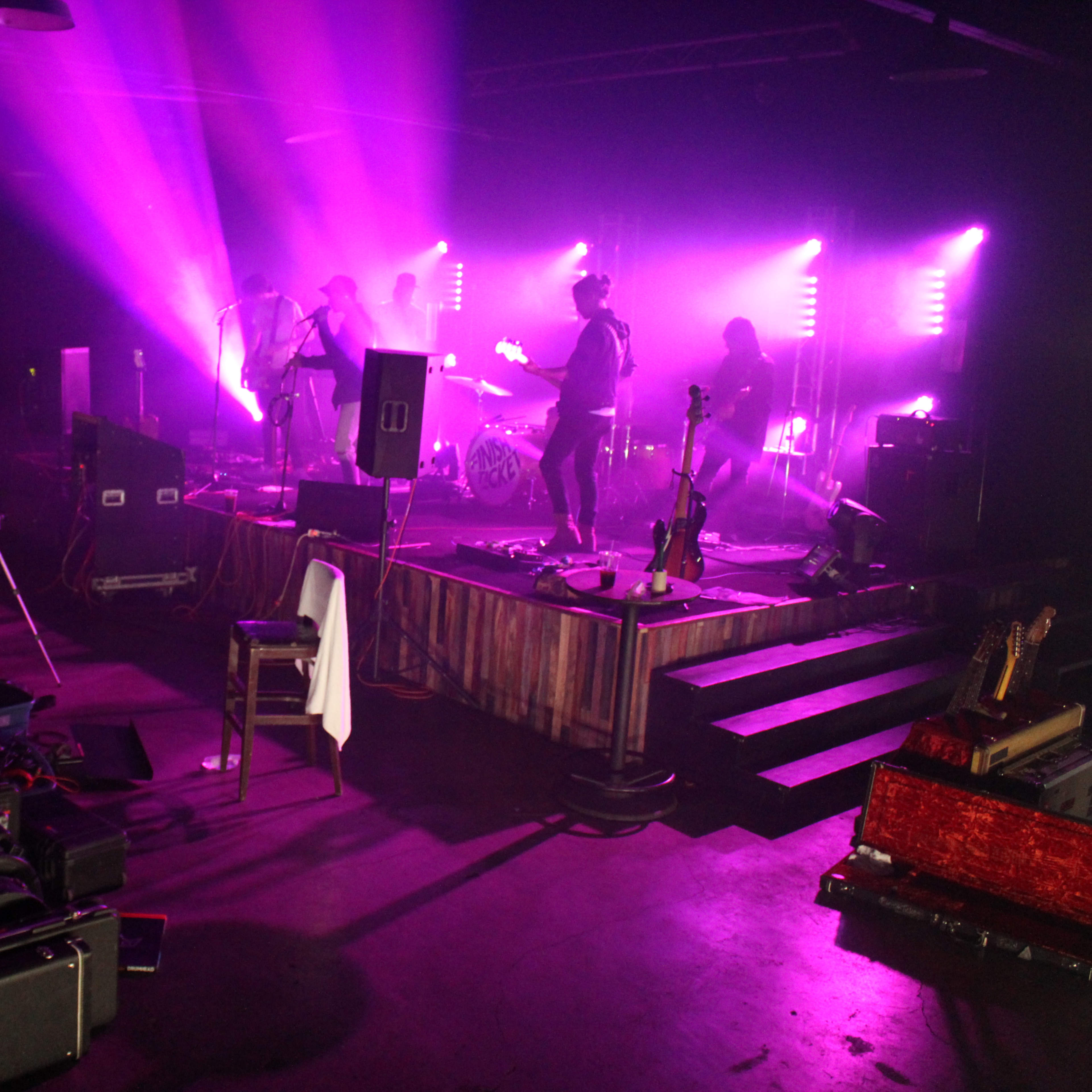 A band performs on stage with purple lighting in a dimly lit venue surrounded by musical equipment.