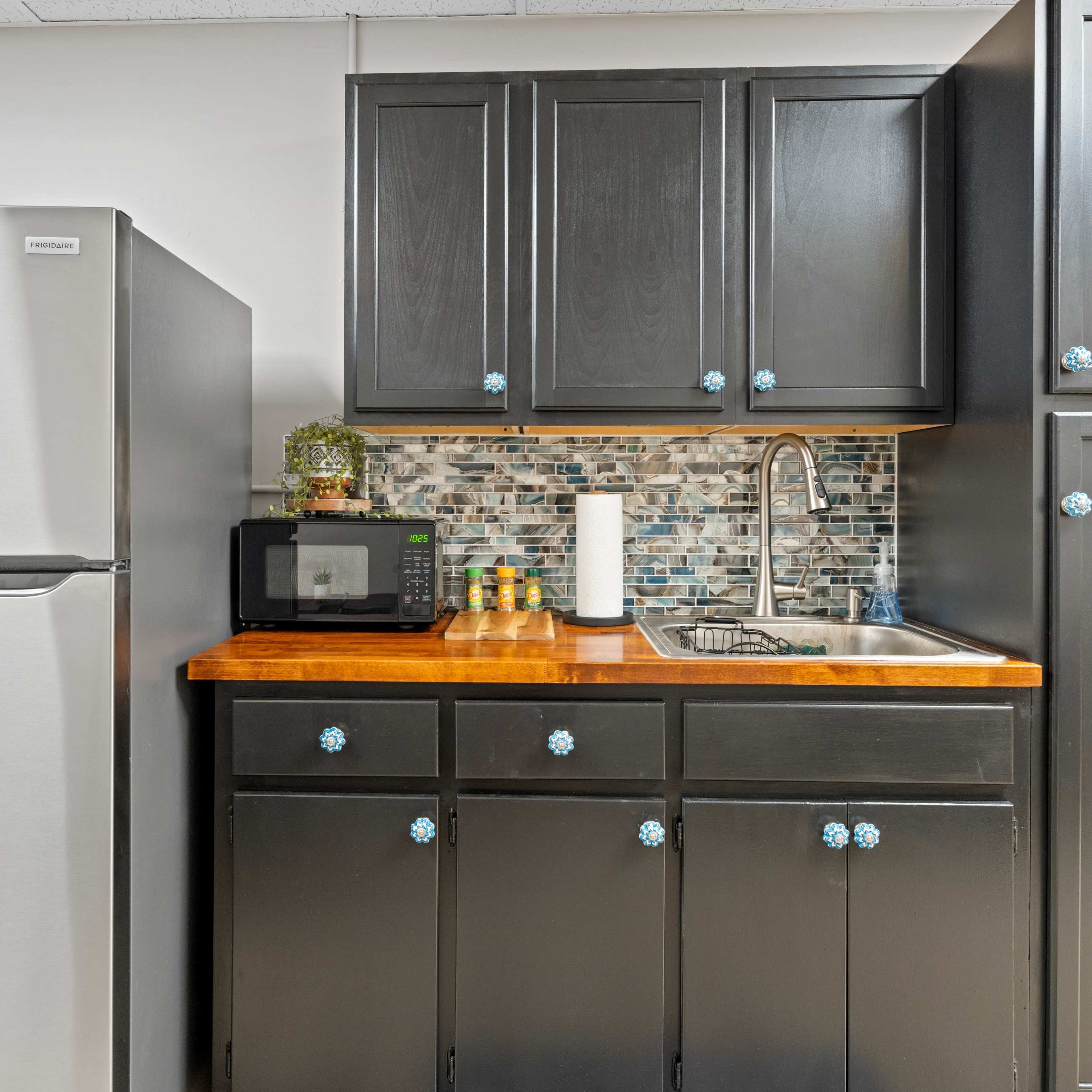 A compact kitchen area featuring a stainless steel refrigerator, a microwave on the countertop, and black cabinets with decorative knobs.