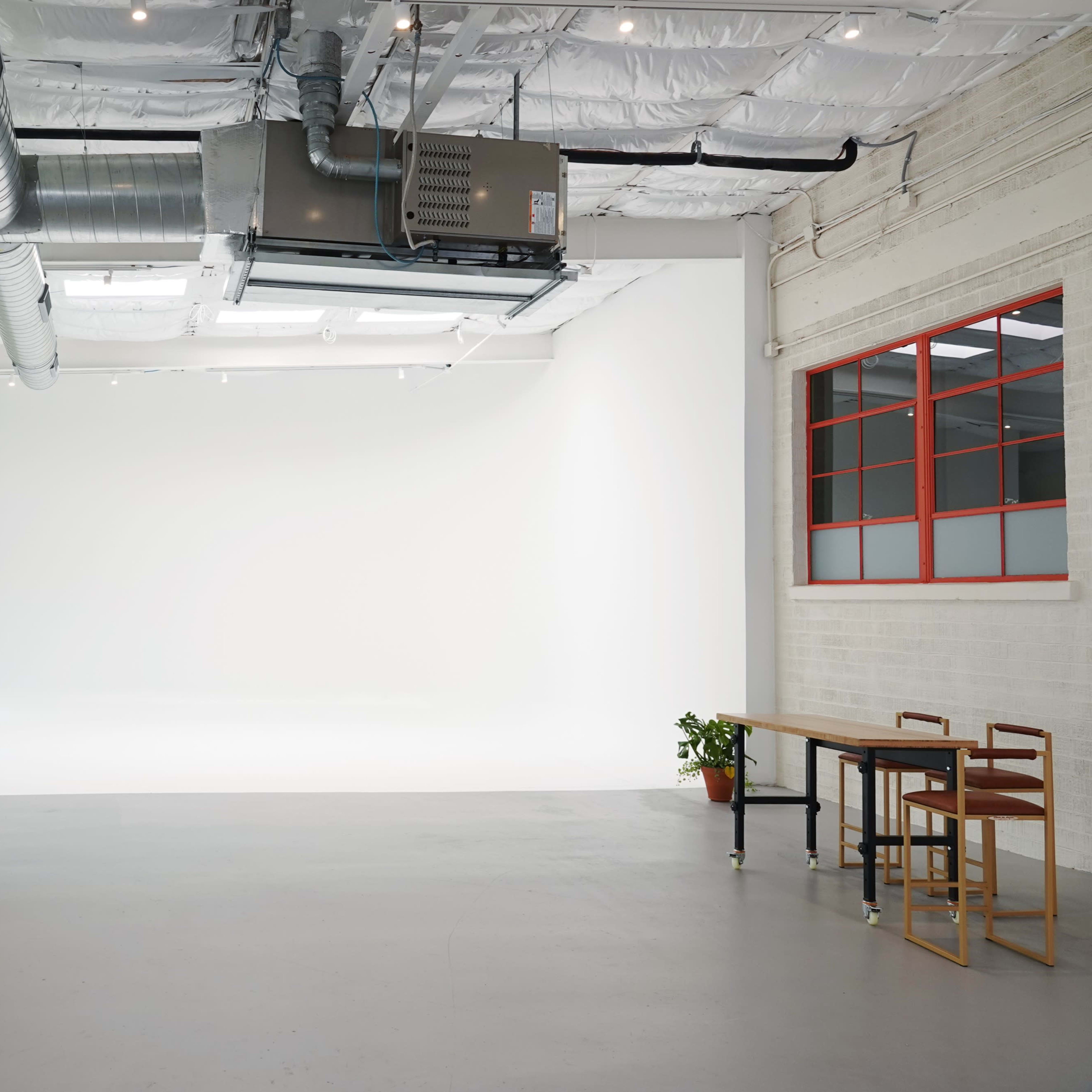 A spacious, well-lit studio with a white backdrop, large windows, and simple furnishings including a table and chairs.