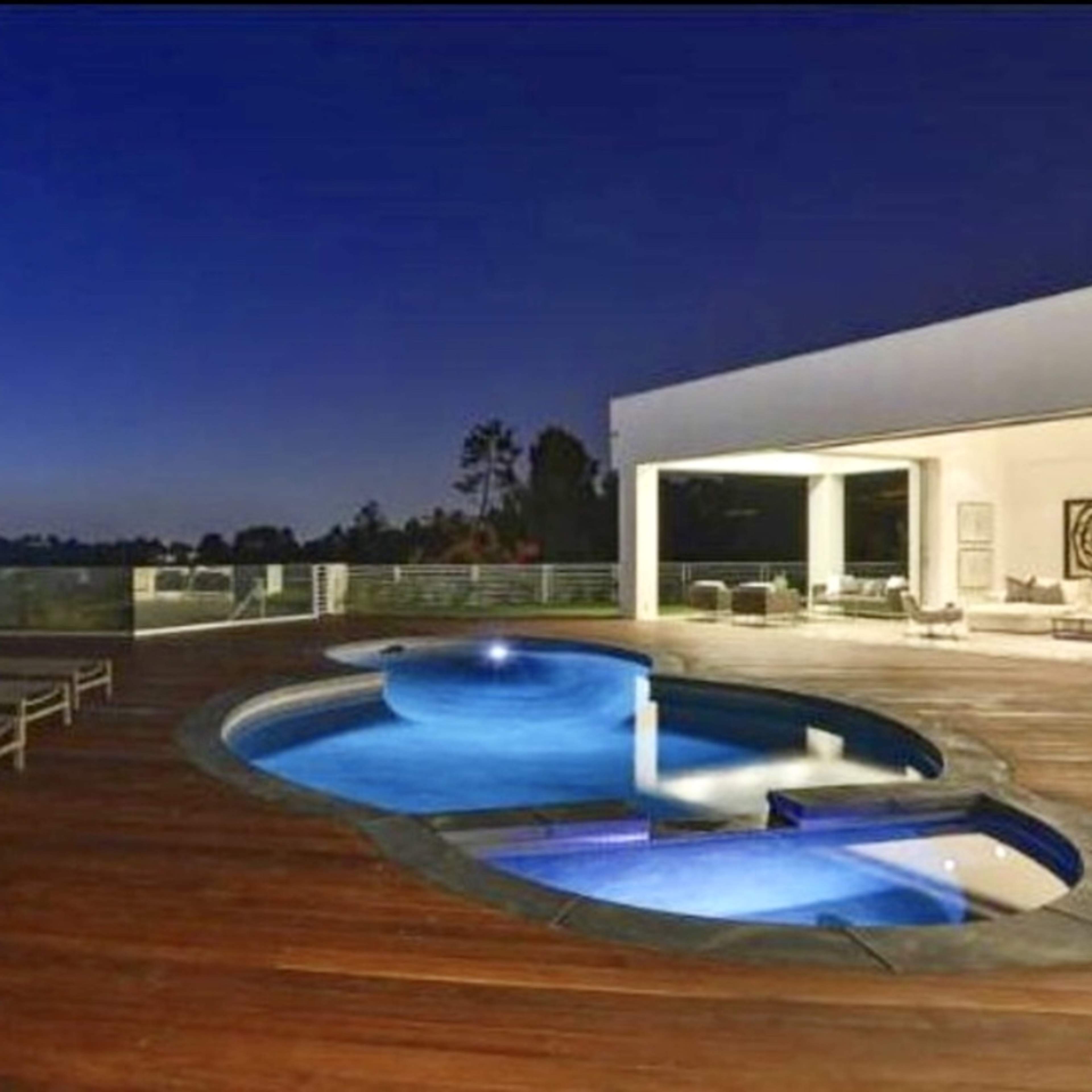 A modern outdoor space featuring a curved swimming pool and lounge chairs on a wooden deck, with a view of the evening sky.