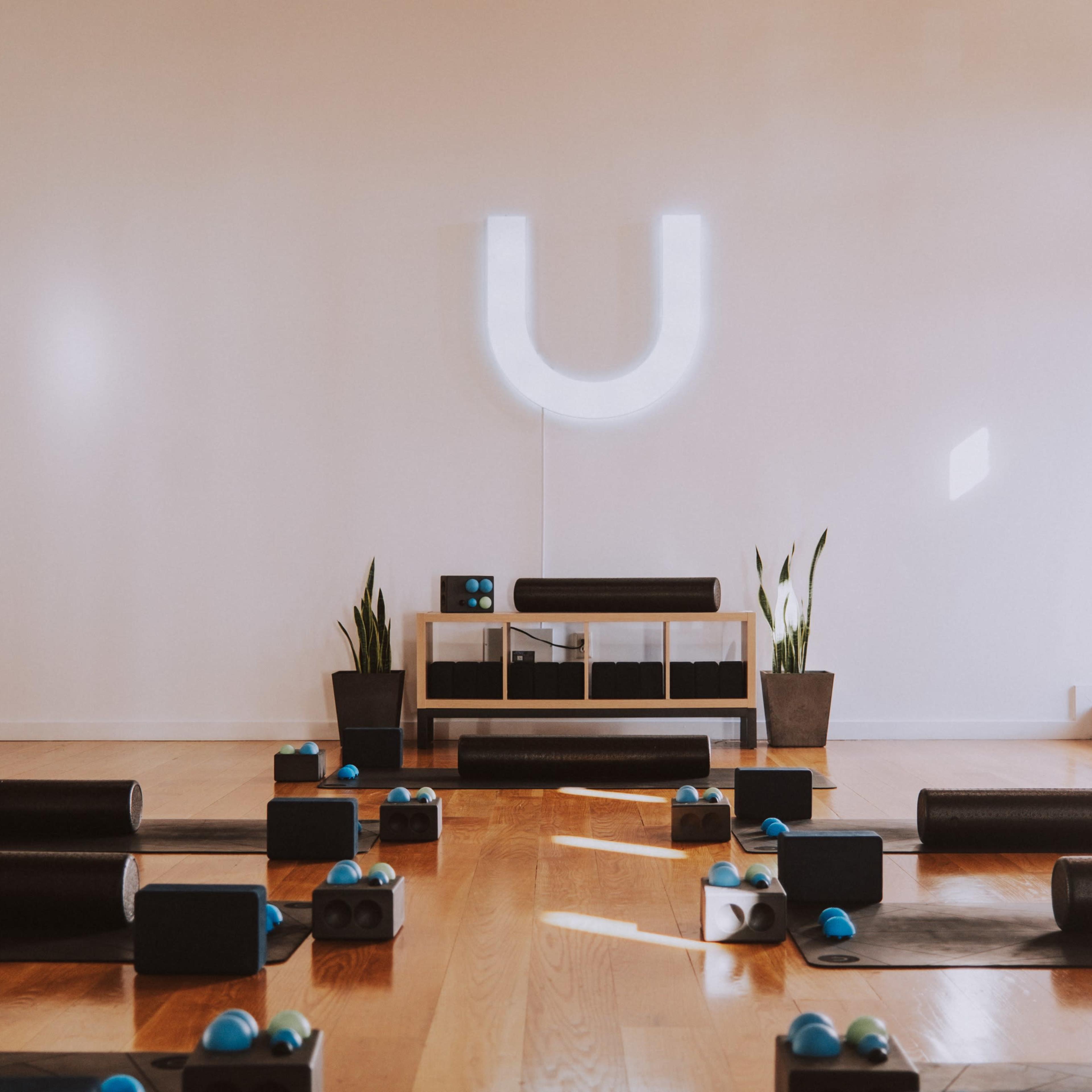 A yoga studio with rolled mats and foam rollers arranged on a wooden floor, featuring a neon sign that says "U" and potted plants in the background.