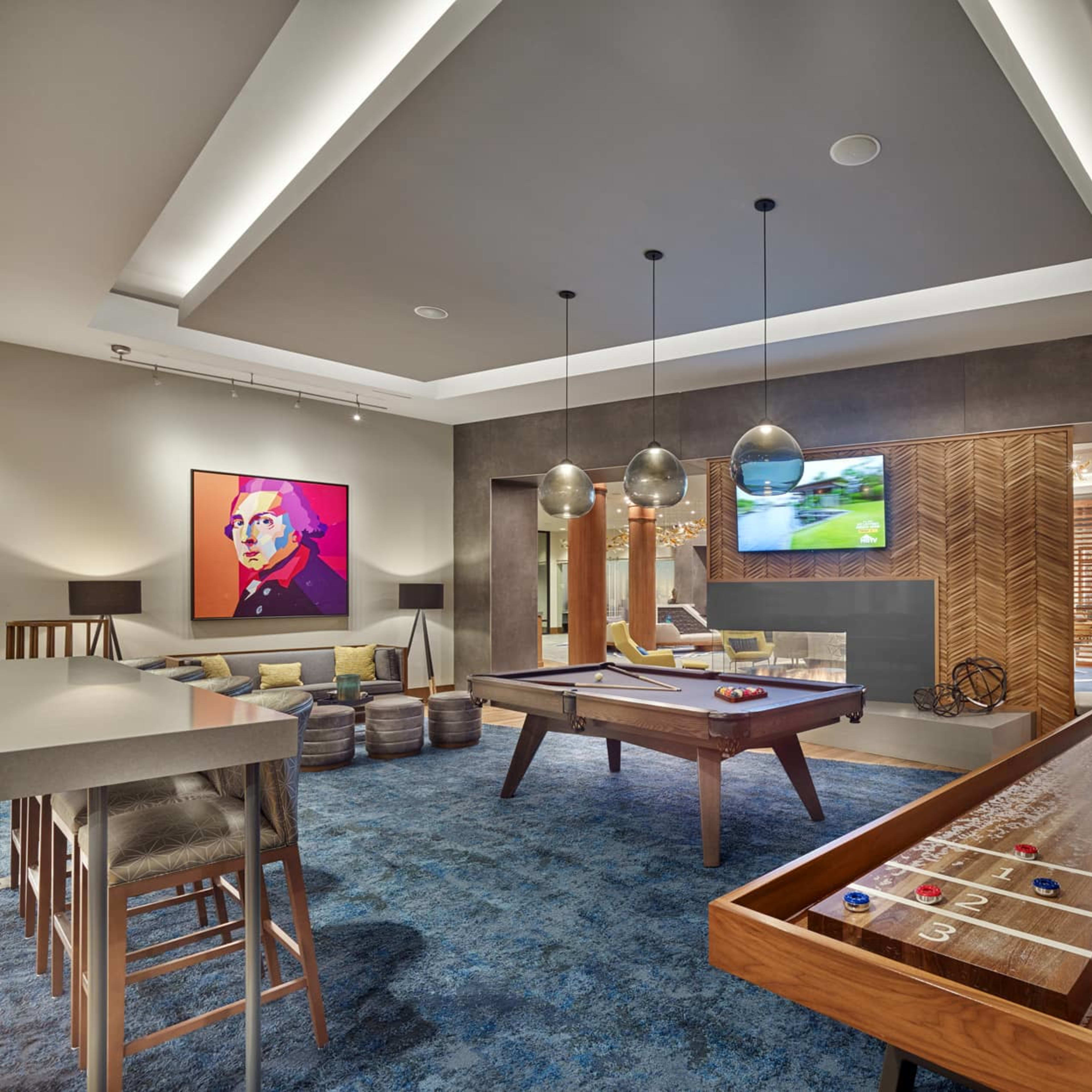 A modern recreation room featuring a pool table, a shuffleboard table, and seating areas, with contemporary decor and lighting.