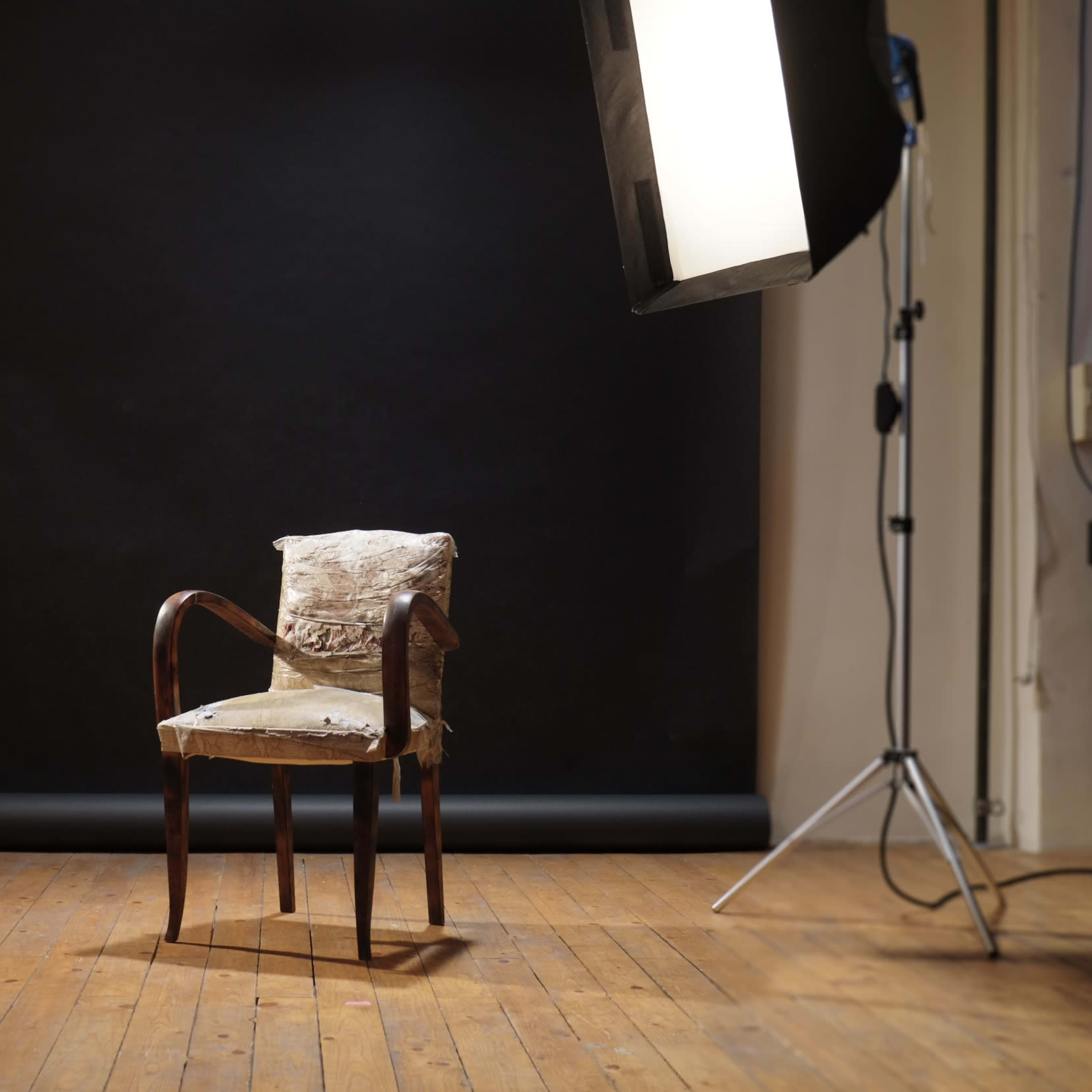 A worn chair is placed in front of a black backdrop, illuminated by studio lights on either side.