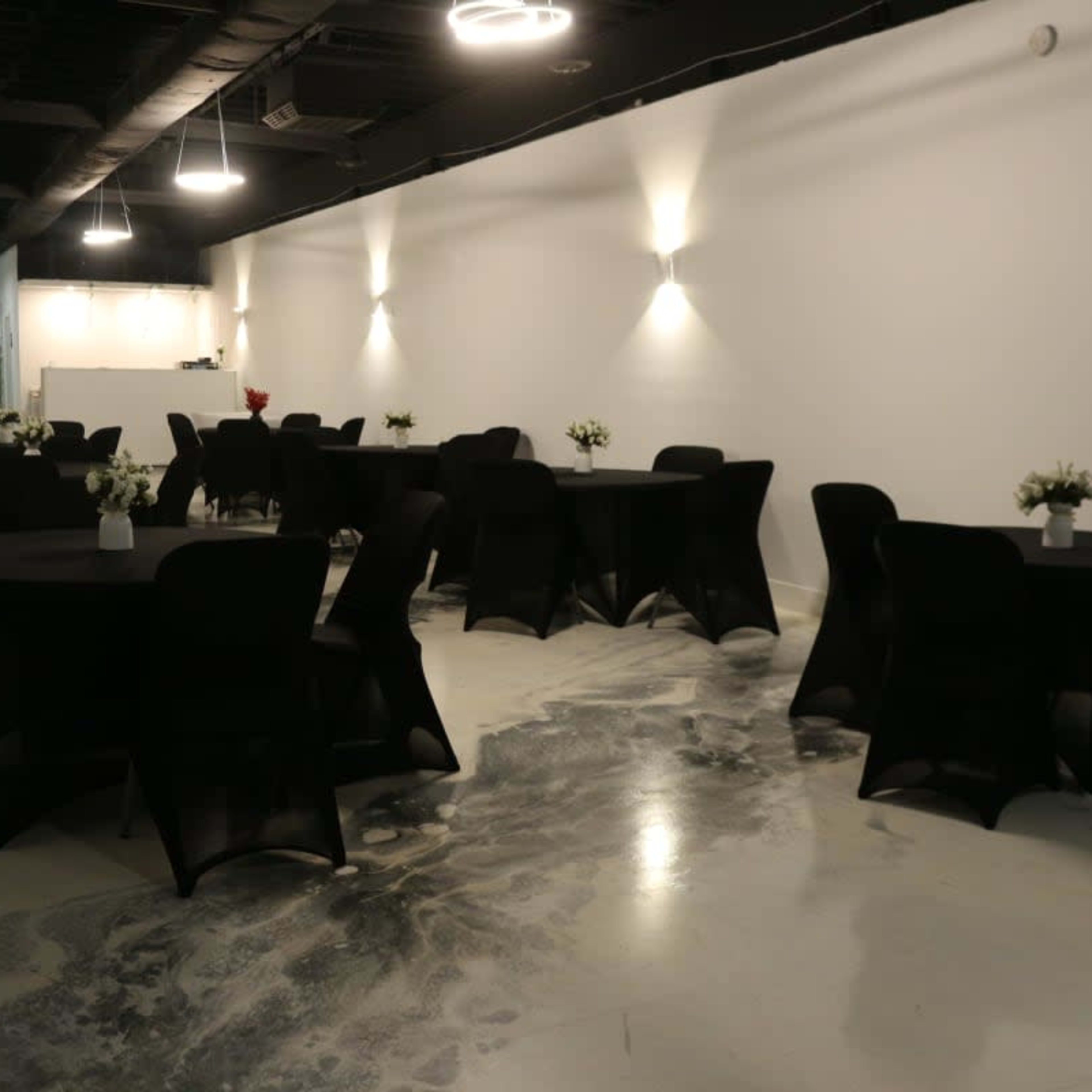 A modern event space with multiple round black tables set with small flower centerpieces, illuminated by wall-mounted lights.