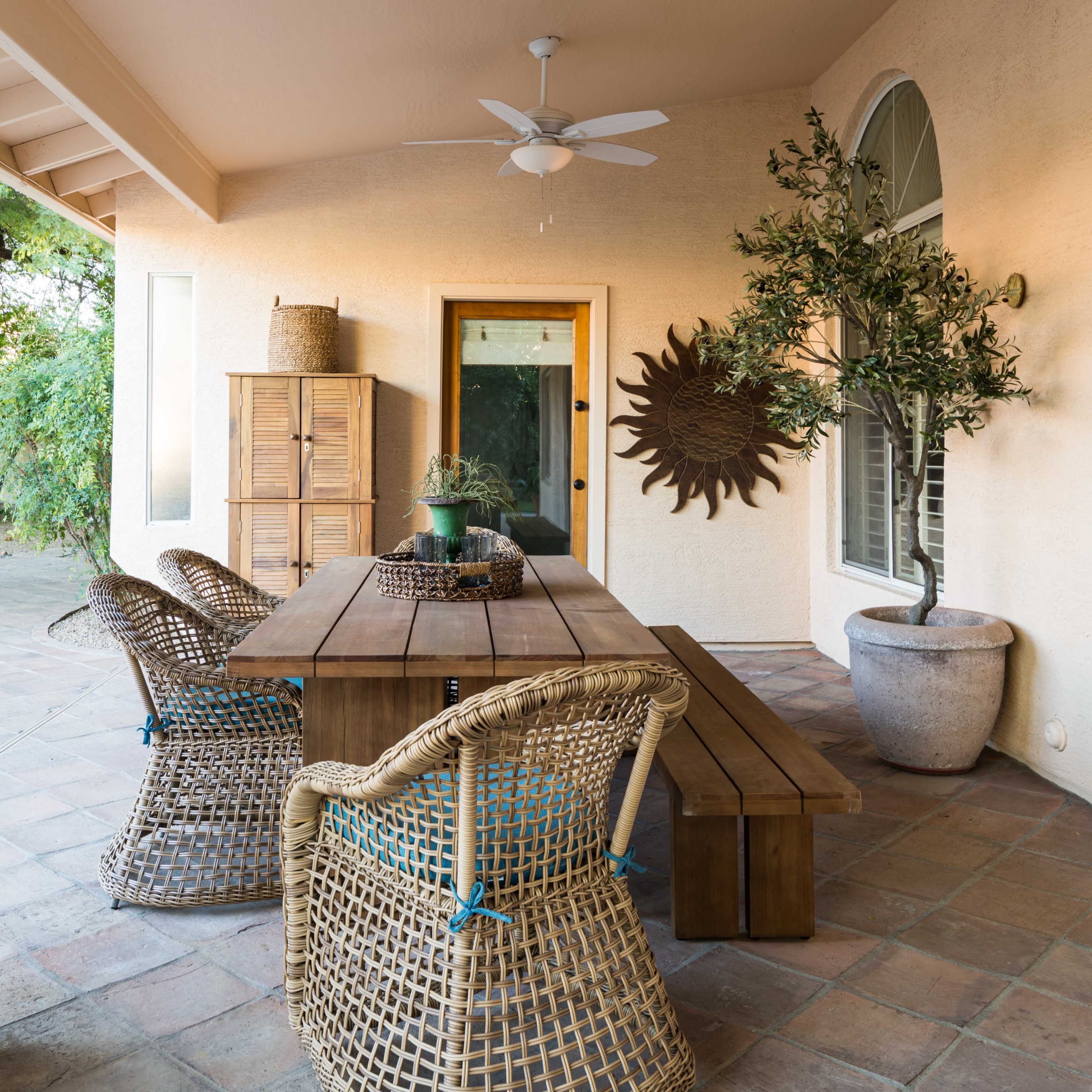 A covered patio features a wooden dining table surrounded by woven chairs, with a potted plant and decorative wall piece nearby.