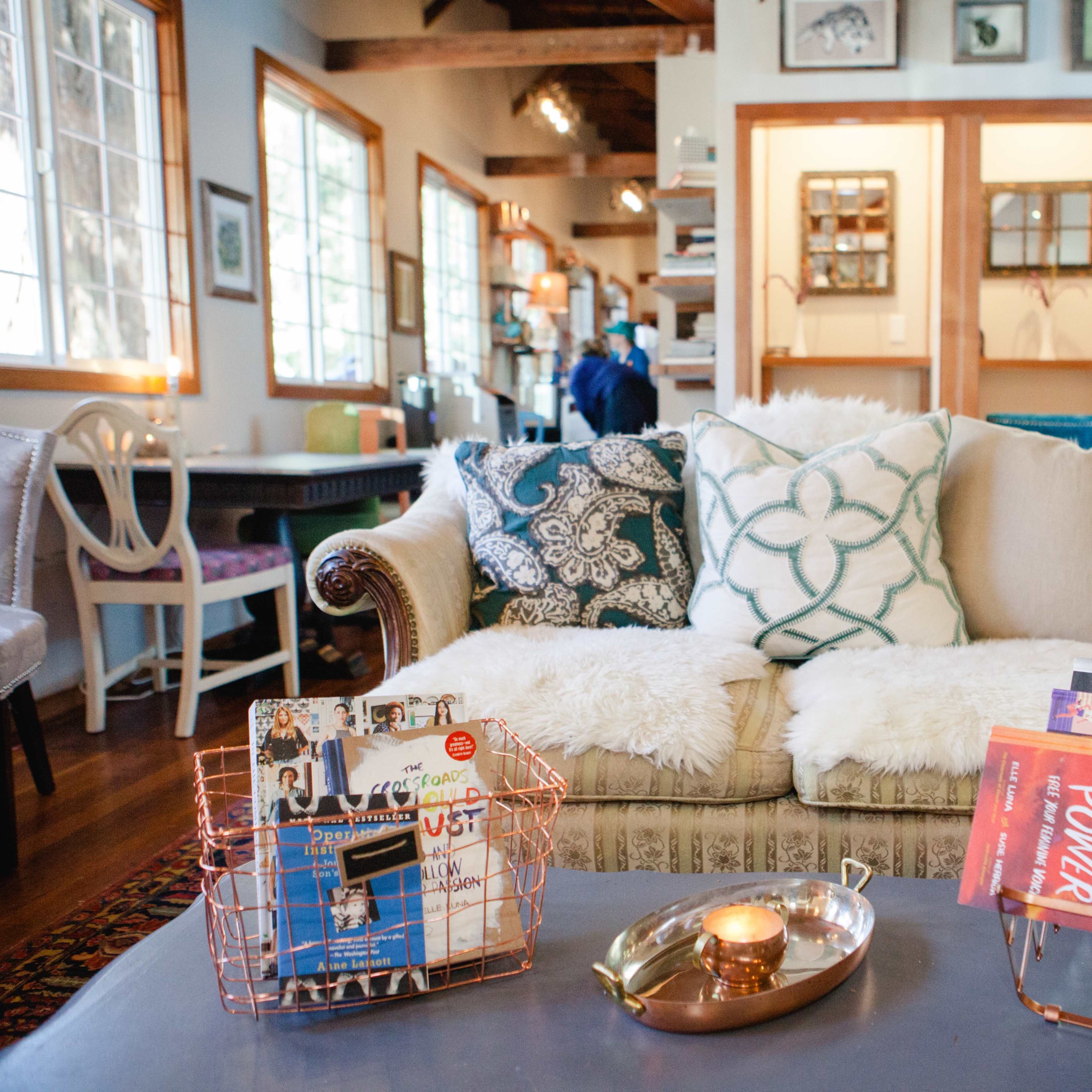 A cozy living room with a patterned sofa, a coffee table displaying books and a candle, and large windows allowing natural light to illuminate the space.