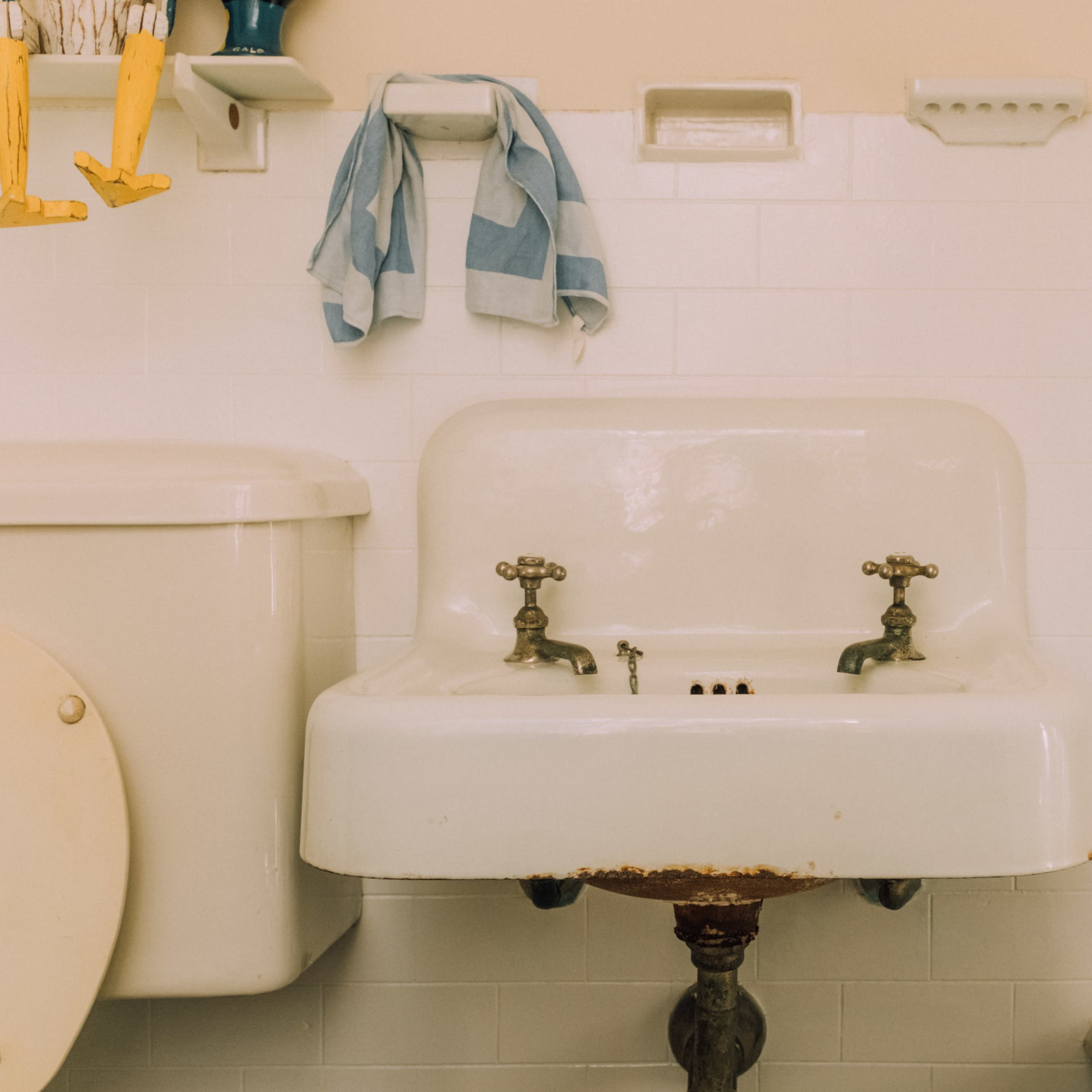 A white porcelain sink with two faucets mounted on a tiled bathroom wall, alongside a toilet and a towel hanging nearby.