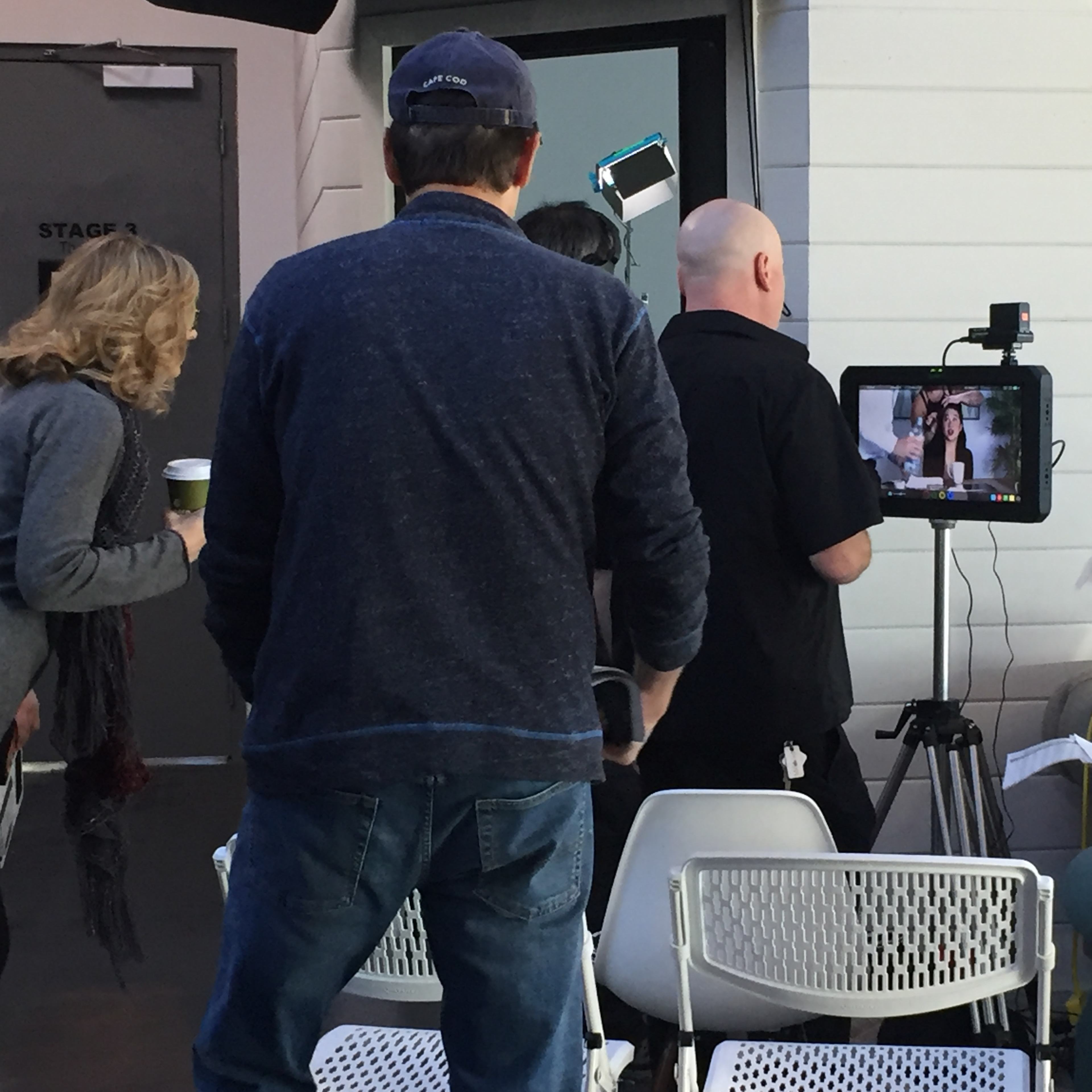 A group of people is gathered around a camera setup on a tripod, focusing on a woman speaking into a microphone.