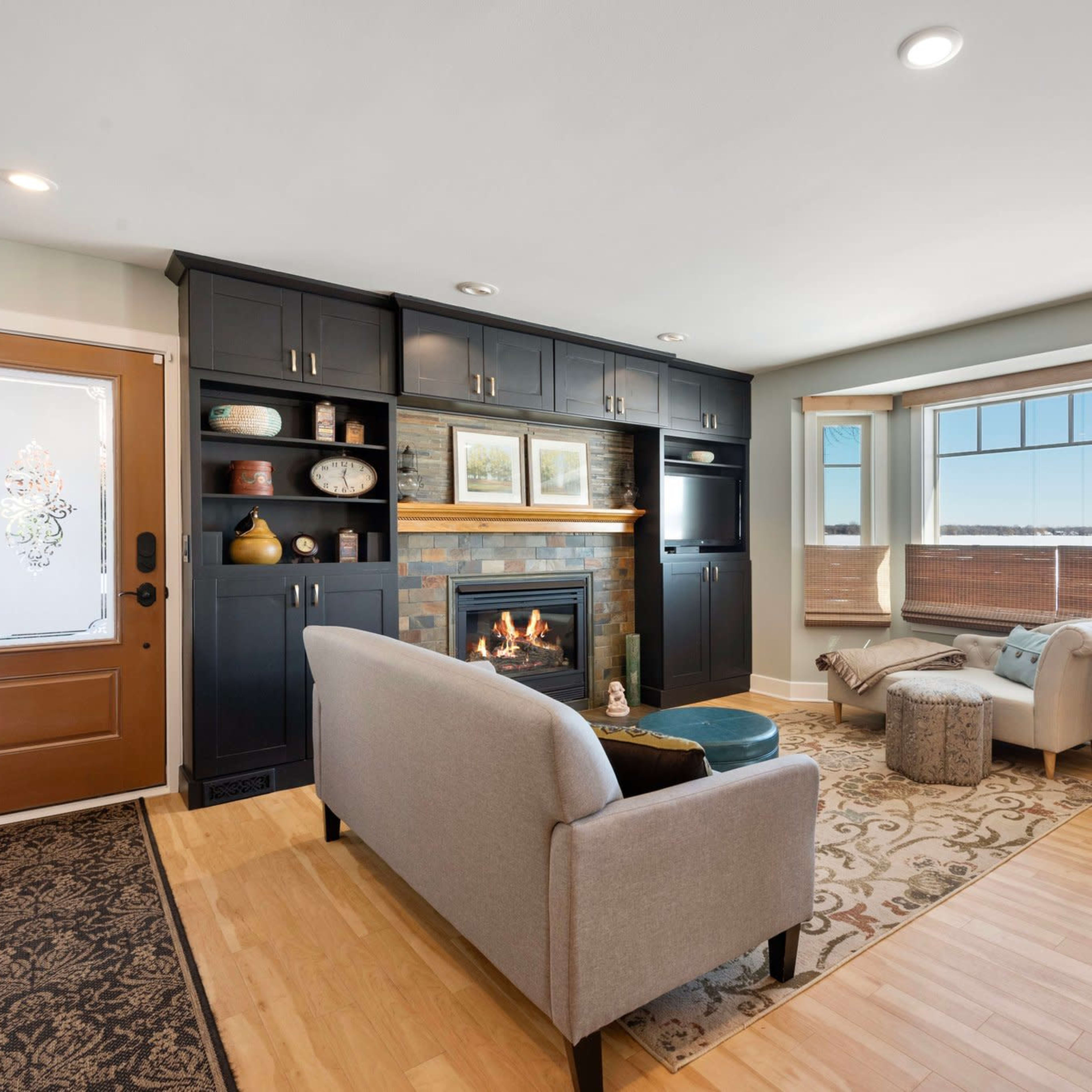 A cozy living room features a fireplace, natural light from large windows, and modern furnishings including a gray sofa and a decorative armchair.