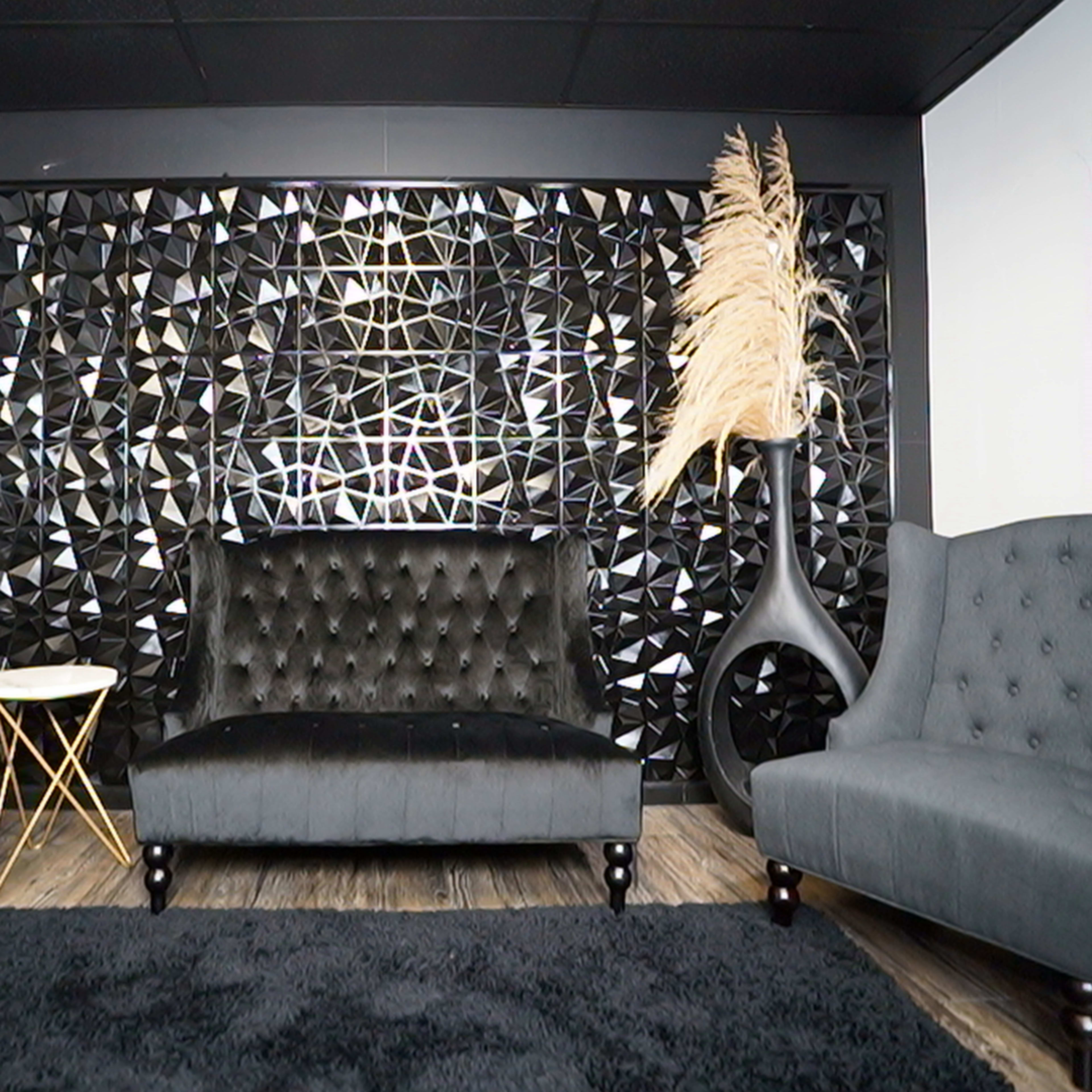 A modern living space with two gray armchairs flanking a black tufted couch, set against a geometric patterned wall and a decorative plant.