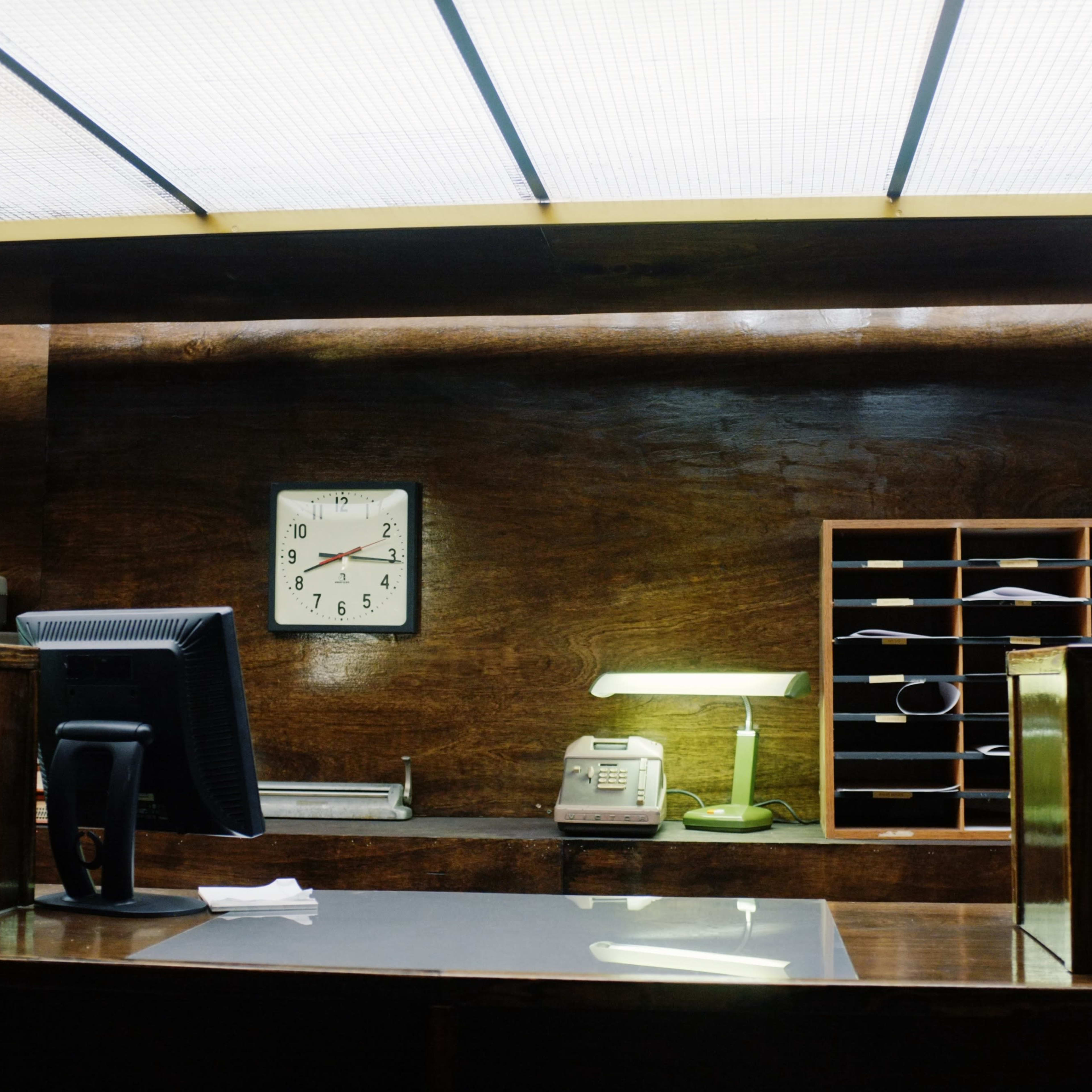 A wooden reception desk with a clock, computer monitor, phone, and an organized mail slot in a dimly lit office.