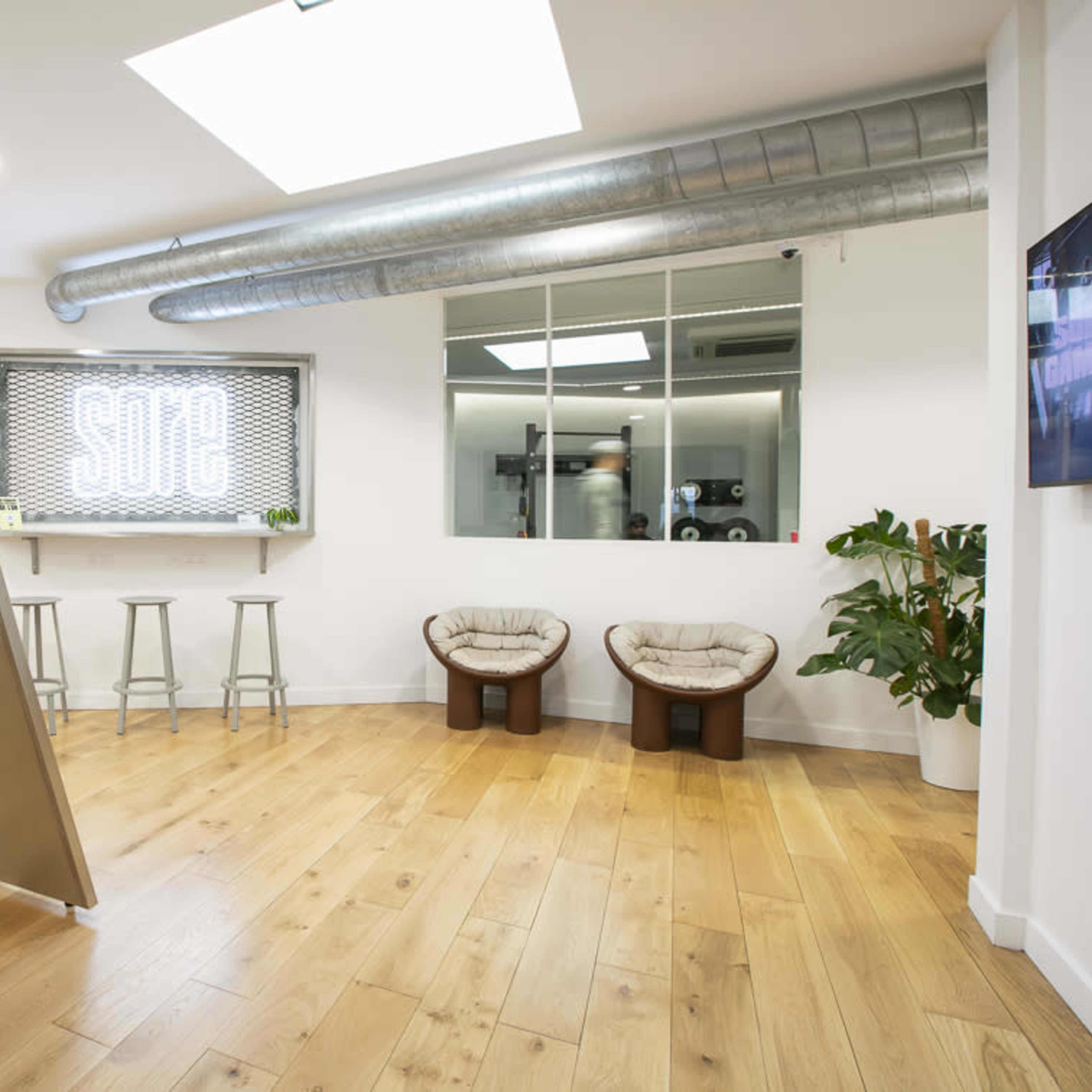 A modern office space features wooden flooring, a metal table with stools, and two plush chairs next to a large window displaying the word "SODE."