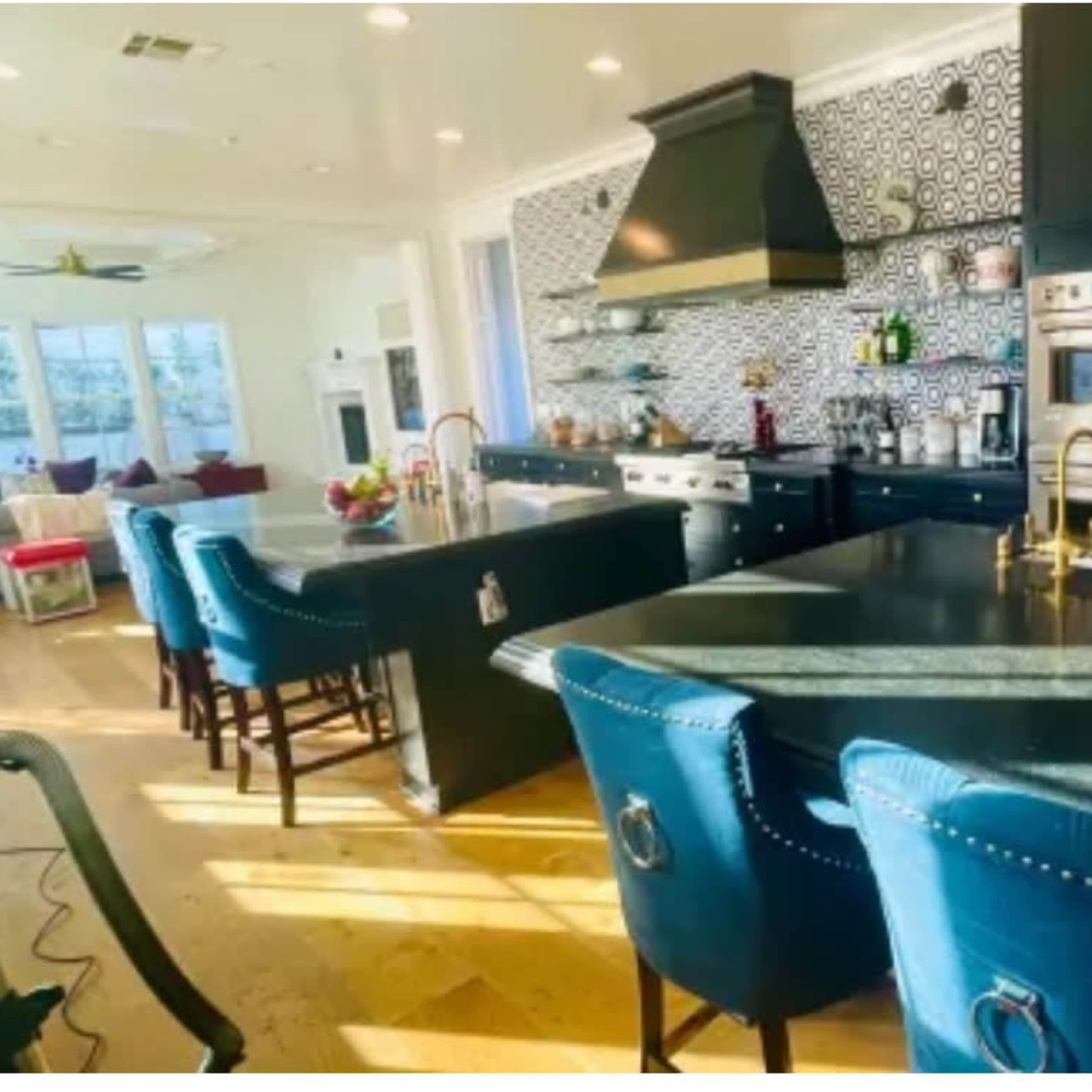 A modern kitchen with dark cabinetry, a black countertop, and a backsplash featuring geometric patterns, illuminated by natural light streaming through large windows.