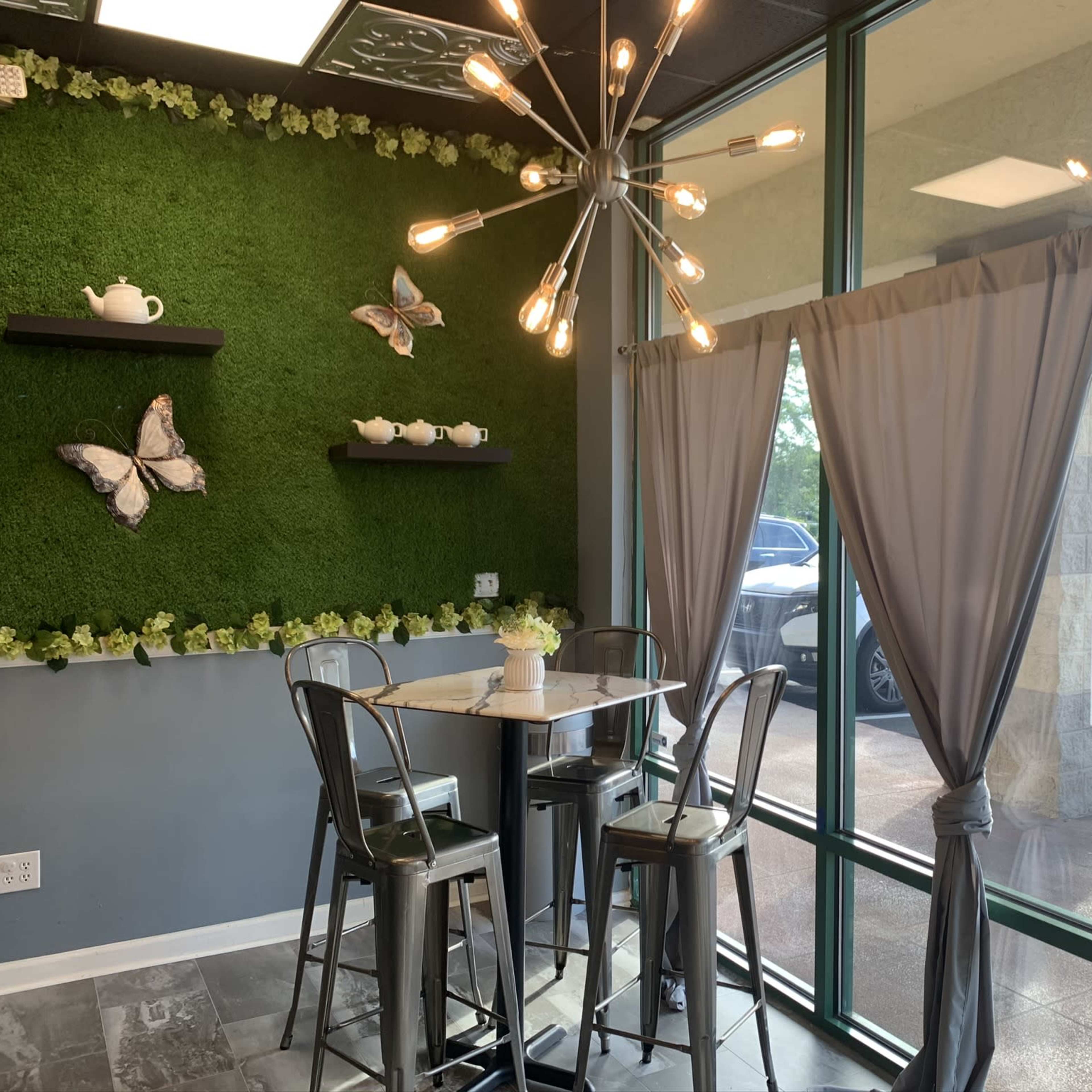 A small cafe interior featuring a green moss wall with decorative butterflies, a table with metal stools, and a large glass entrance with sheer curtains.