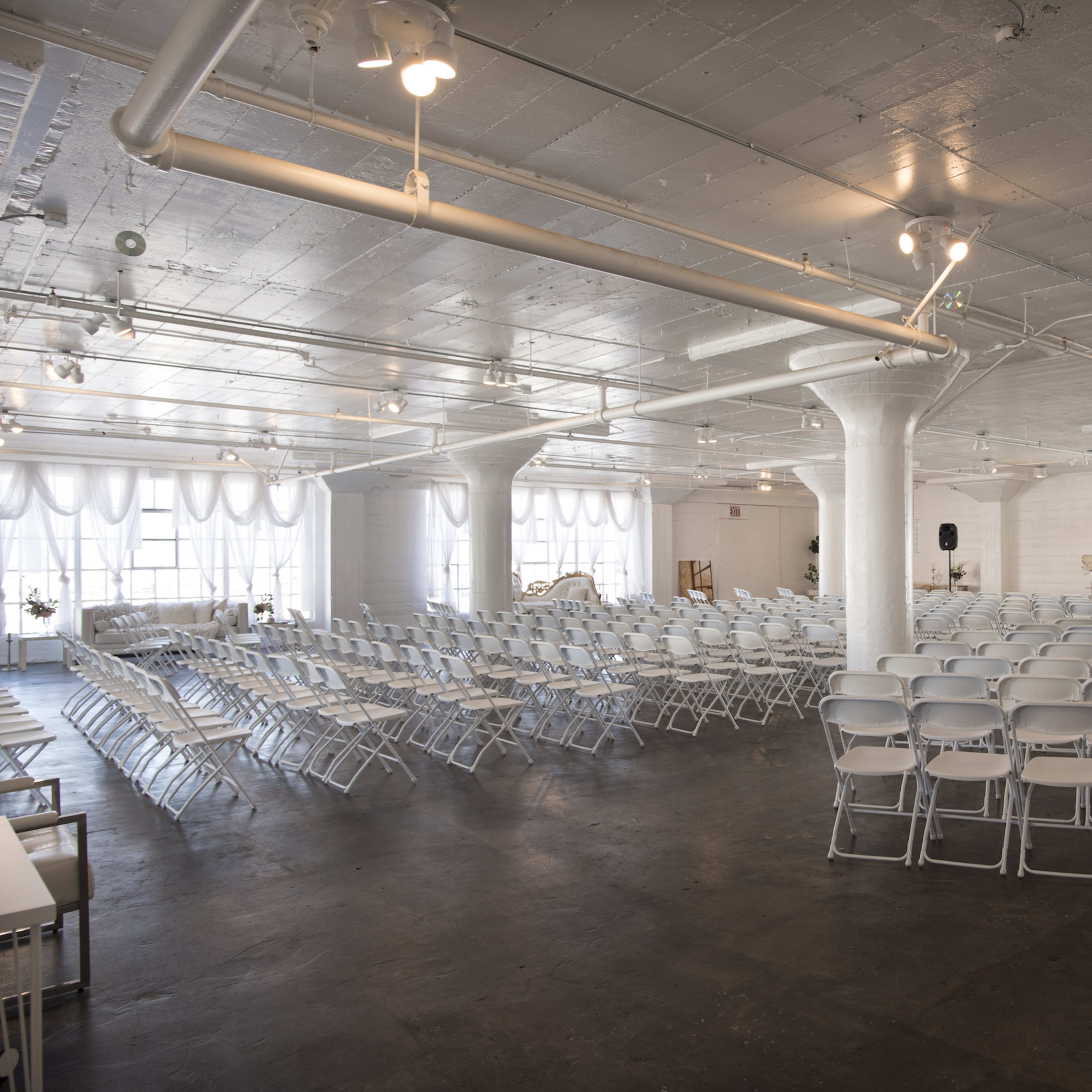 A spacious, well-lit event room arranged with rows of white folding chairs facing a stage area.