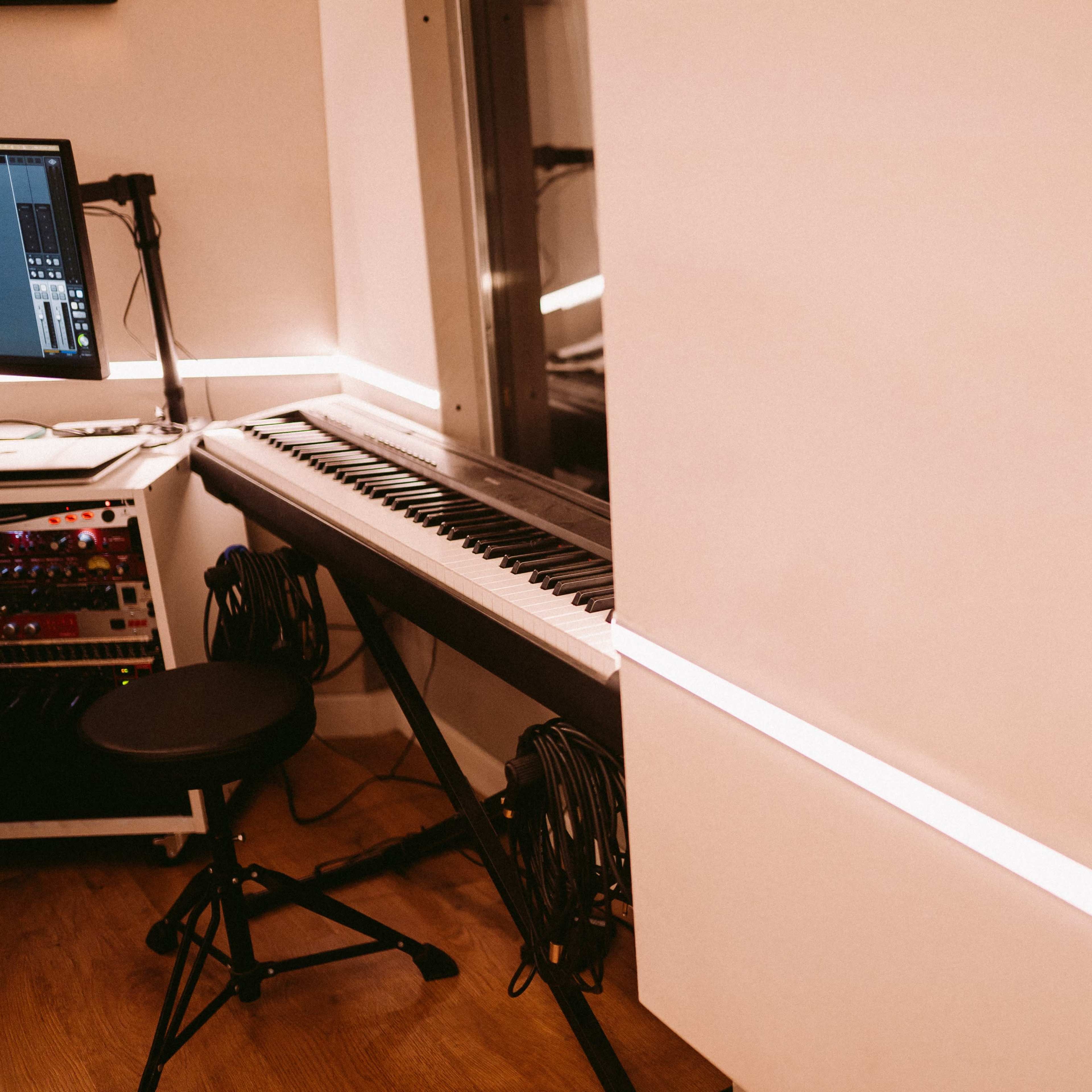A digital piano is positioned next to a computer setup in a music studio, with a chair and equipment visible in the background.