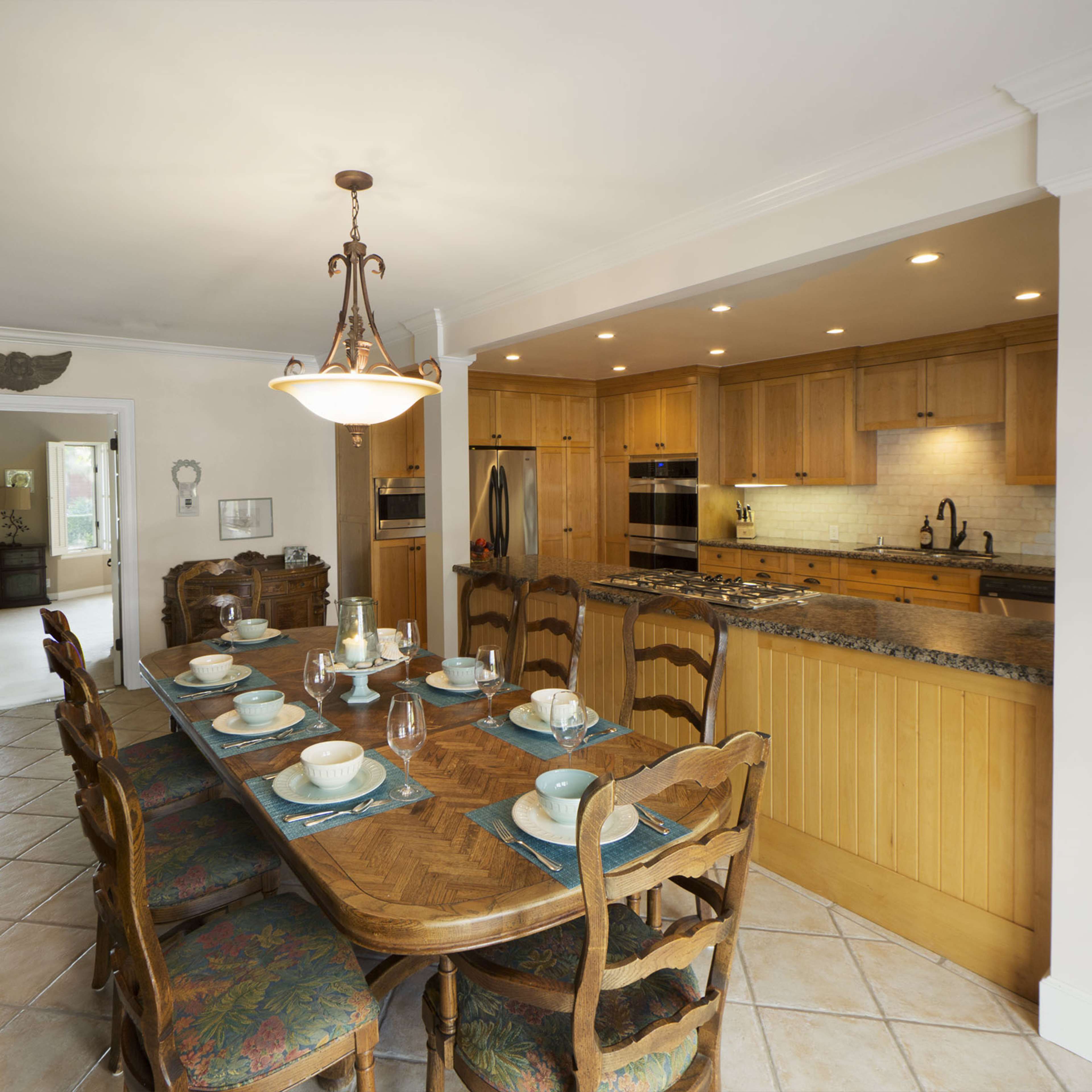 A large dining table is set for a meal in a warm-toned kitchen with wooden cabinetry and modern appliances.